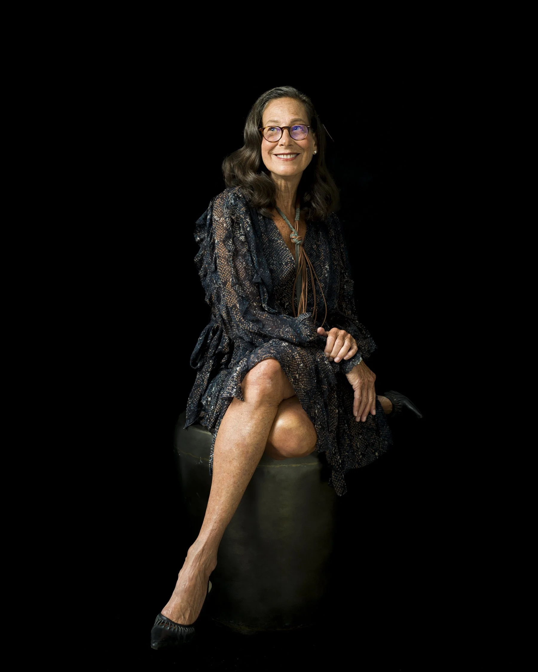 A woman with long brown hair, glasses, and a patterned dress sitting on a stool with her legs crossed, smiling against a black background.
