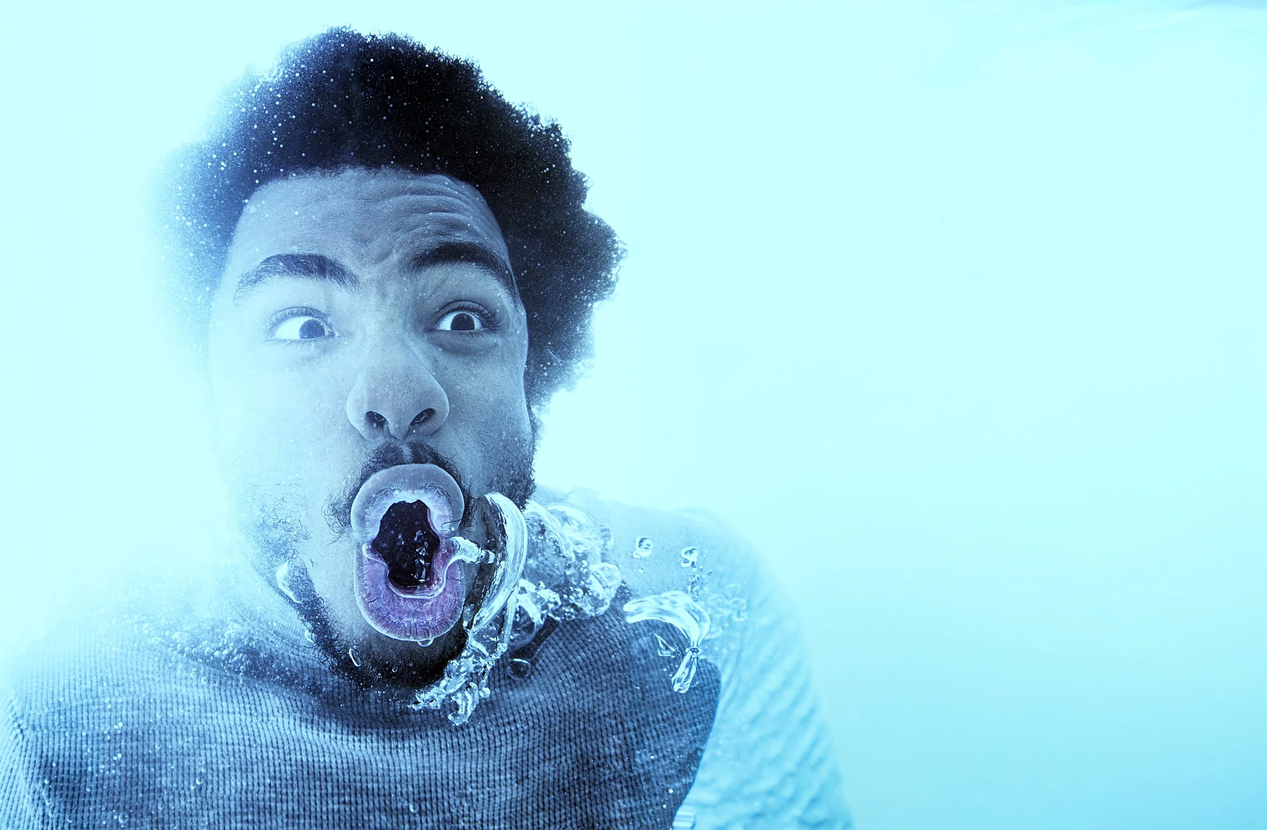A man with curly hair and a beard underwater, with bubbles around his face, making a surprised expression with his mouth open.