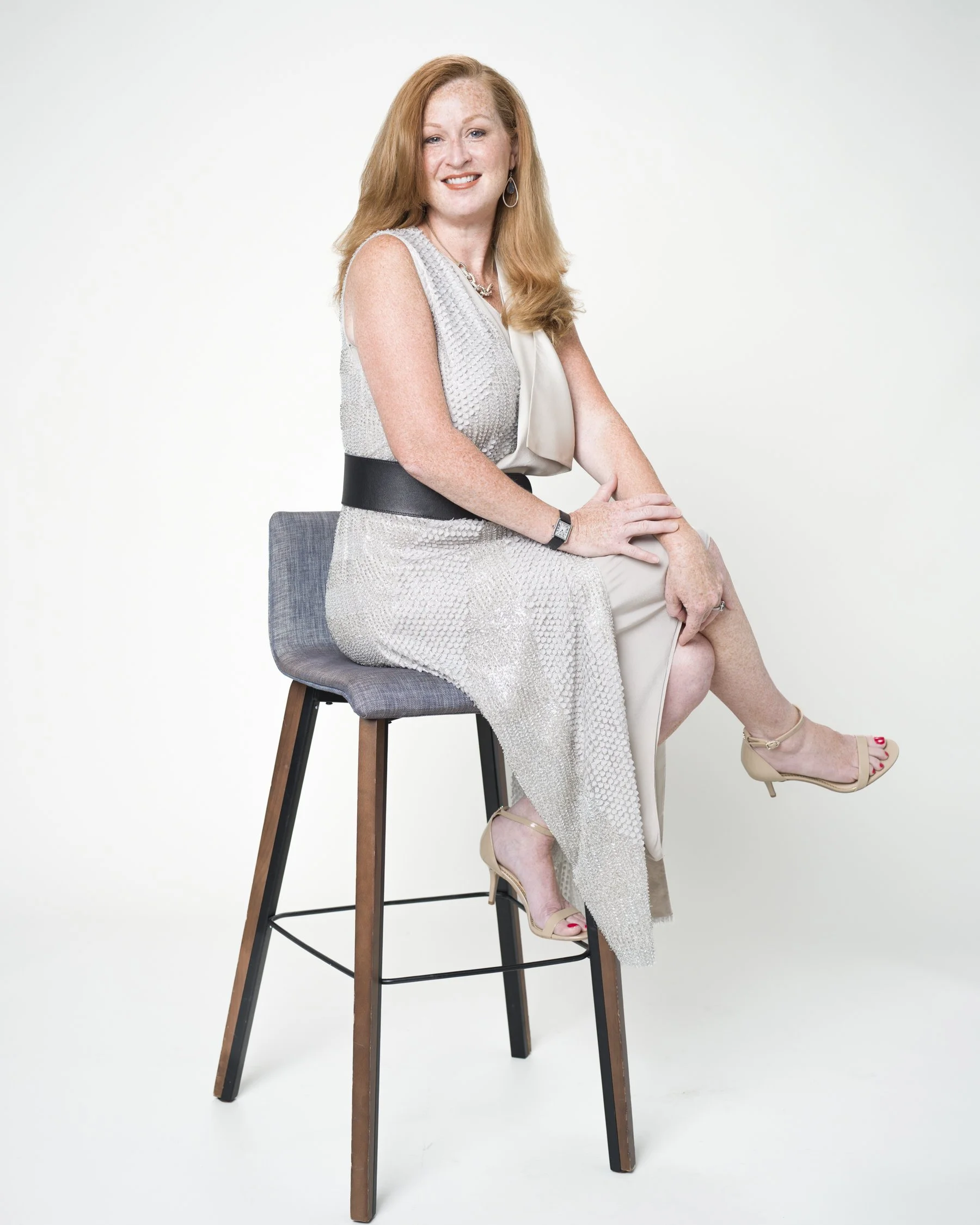 Red-haired woman wearing a white sleeveless top and beige pants, sitting on a tall gray and wood stool against a plain white background.