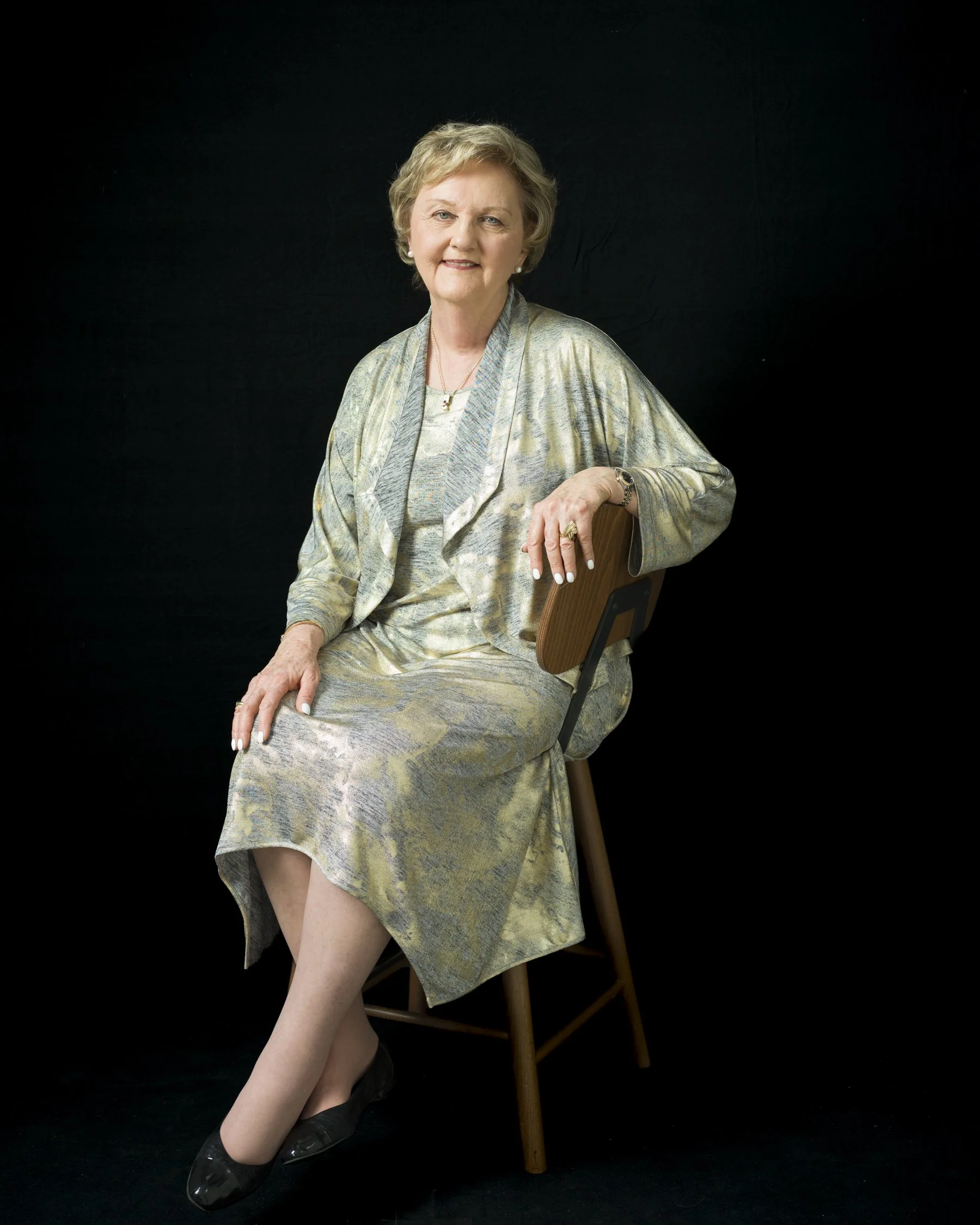 An elderly woman with short blonde hair wearing a light, patterned dress and a matching shawl, sitting on a wooden chair against a black background. She is smiling and looking at the camera, with her right hand resting on her knee and left hand resti