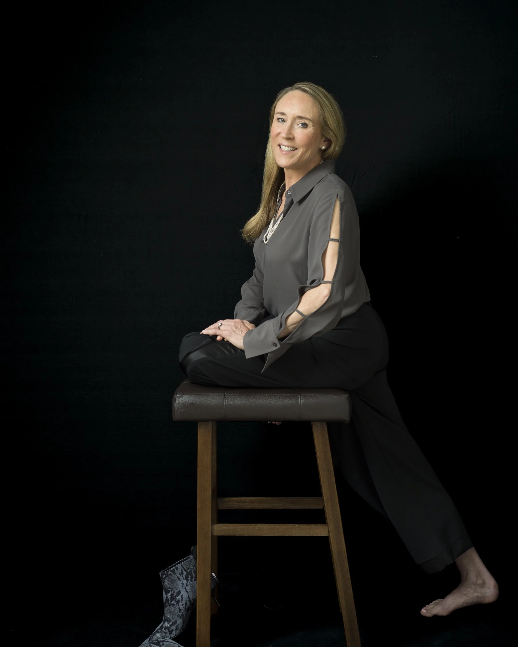 A woman sitting on a wooden chair with her legs crossed at the ankles, wearing a gray blouse, black pants, and patterned boots, smiling against a black background.