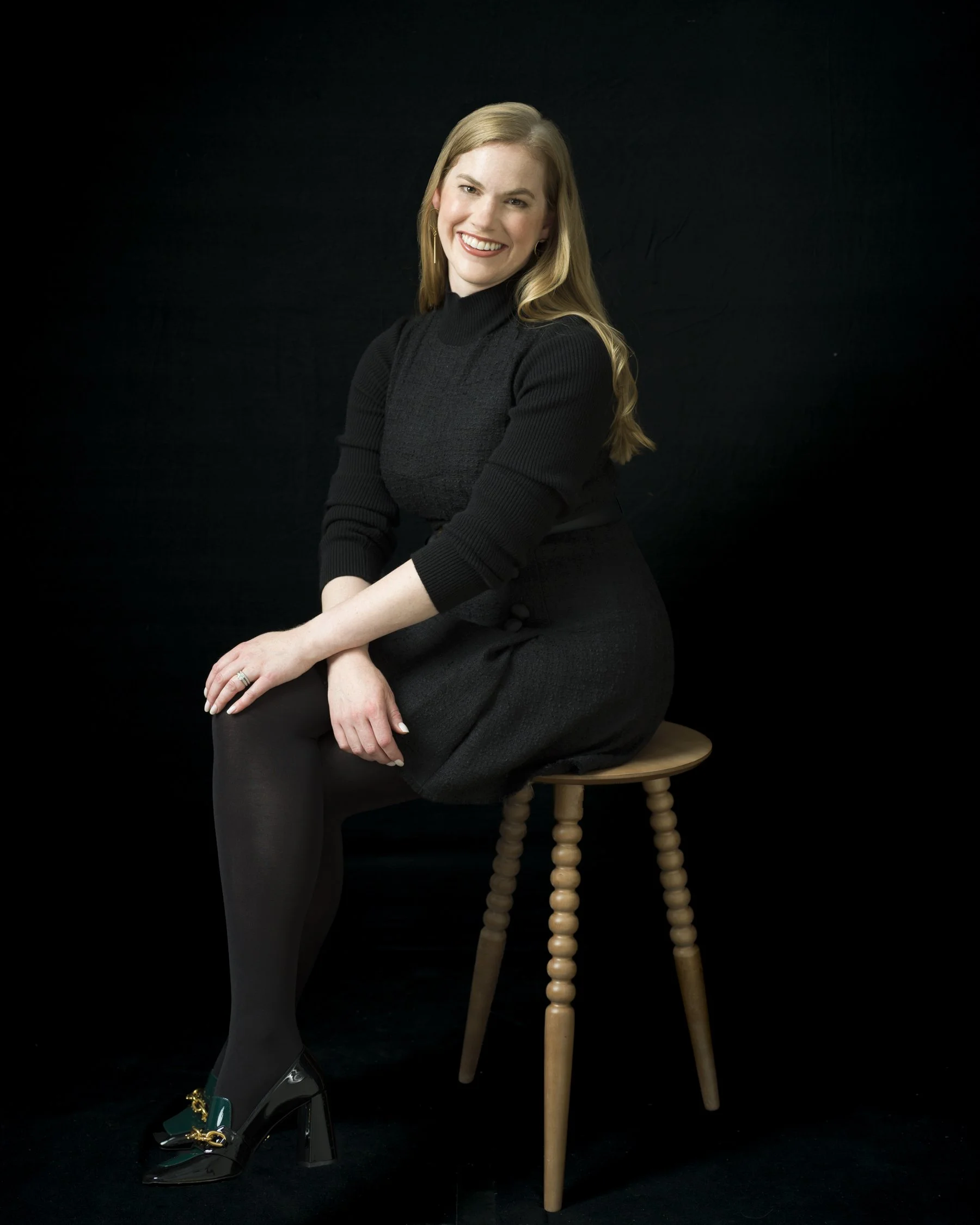 A young woman with long blonde hair smiling, sitting on a wooden stool, dressed in a black turtleneck and black skirt, with one leg crossed over the other, against a black background.