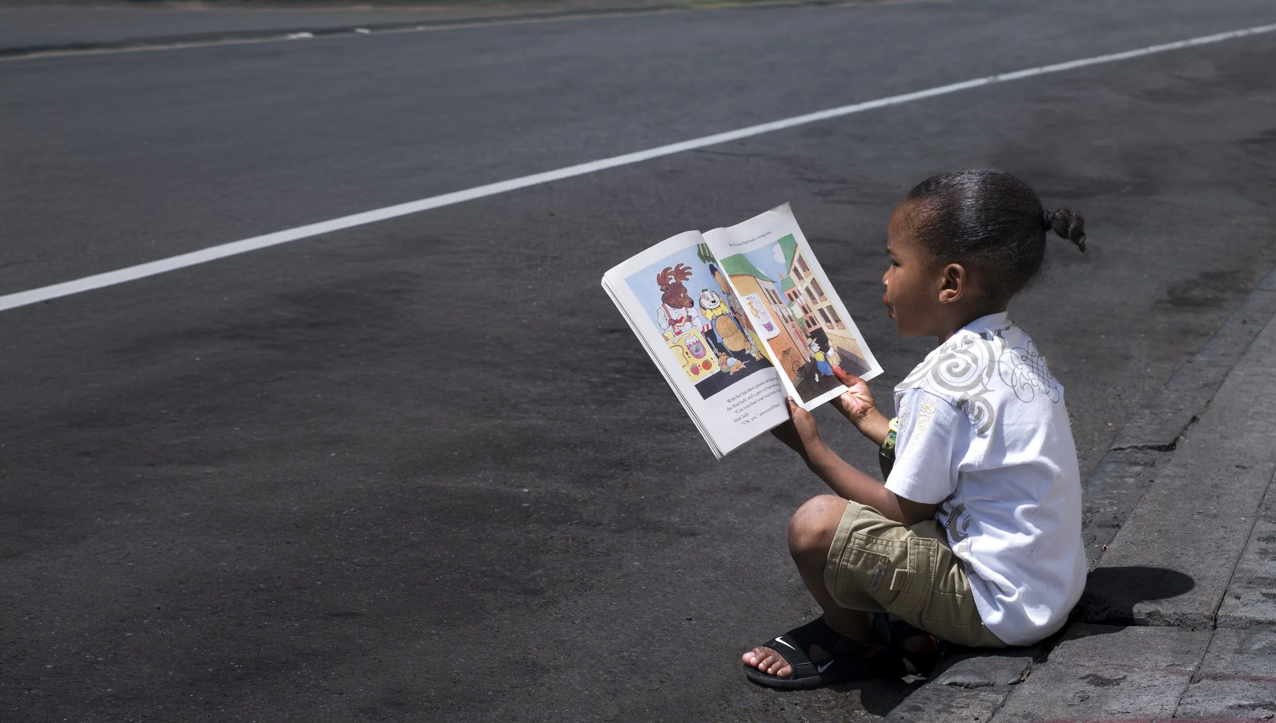 A young boy with dark hair in a bun, wearing a white shirt and khaki shorts, sitting on a sidewalk and reading a colorful picture book with animated characters.