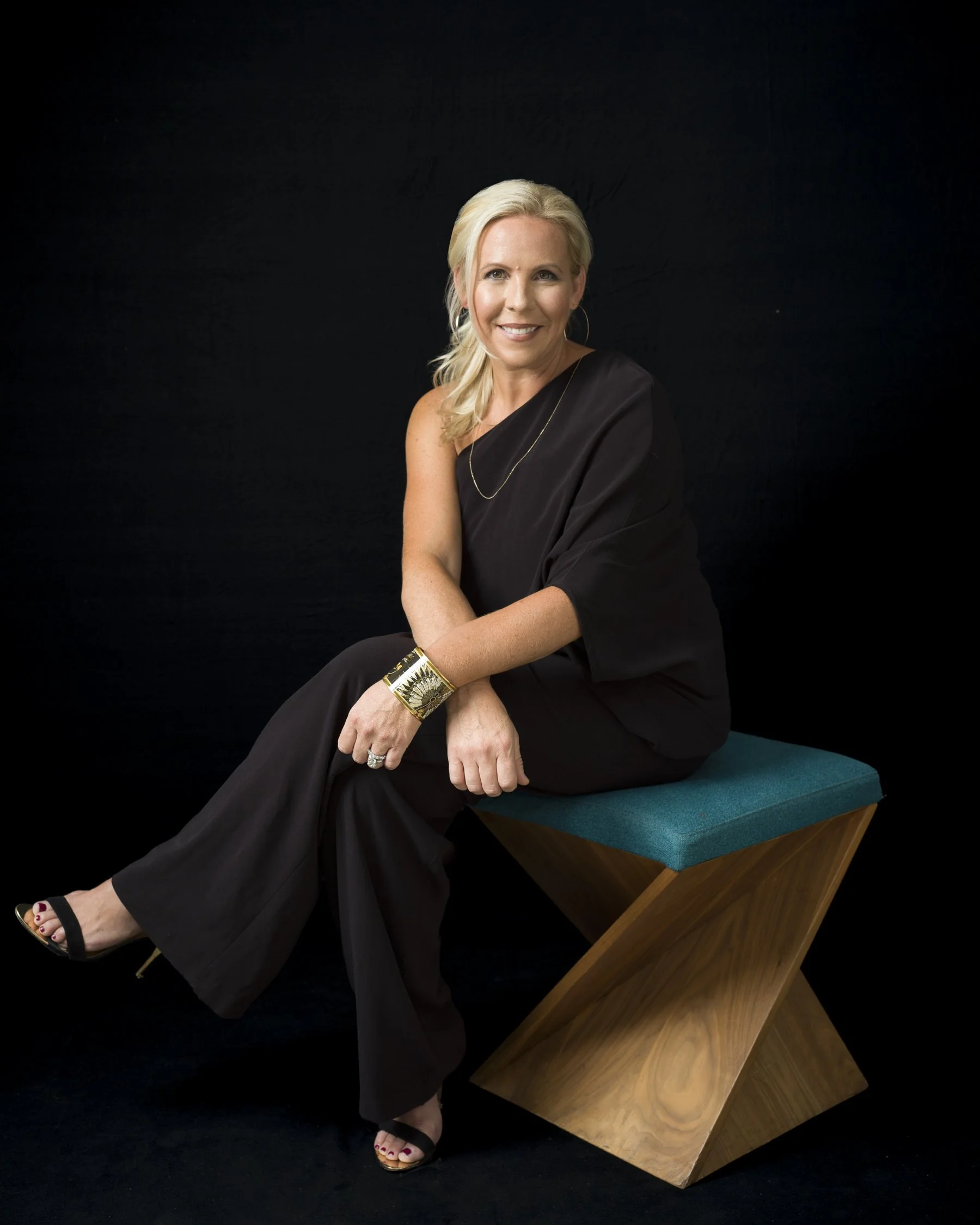 A woman with blonde hair sitting on a teal cushion on a wooden geometric bench, wearing a black dress, gold accessories, and black open-toe heels, against a black background.