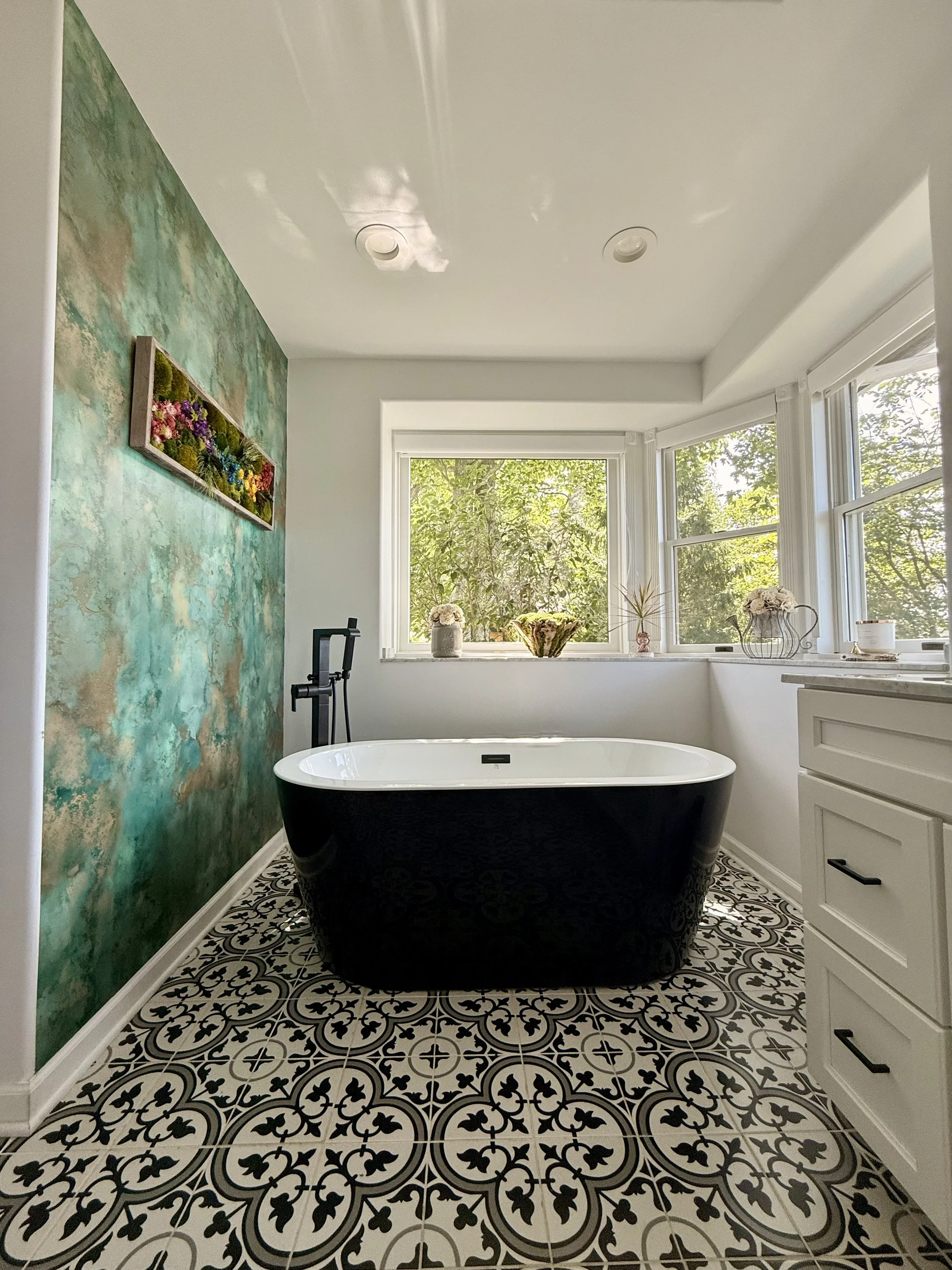 Bathroom after the transformation, featuring a black bathtub, patterned tile floor, and a bold green floral accent wall with artwork, plants, and natural light creating a refreshed and inviting space.