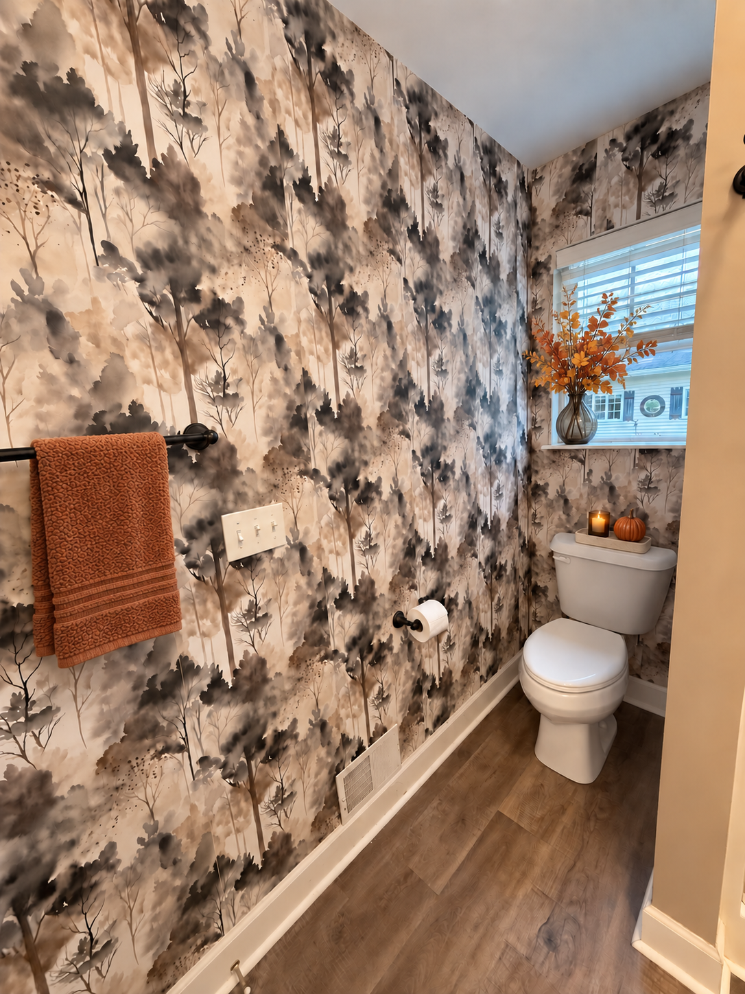 A small bathroom with a black and beige watercolor-themed wallpaper, a white toilet with a pumpkin and candle on top, a window with blinds and a vase of orange flowers, a brown towel, and wooden flooring.
