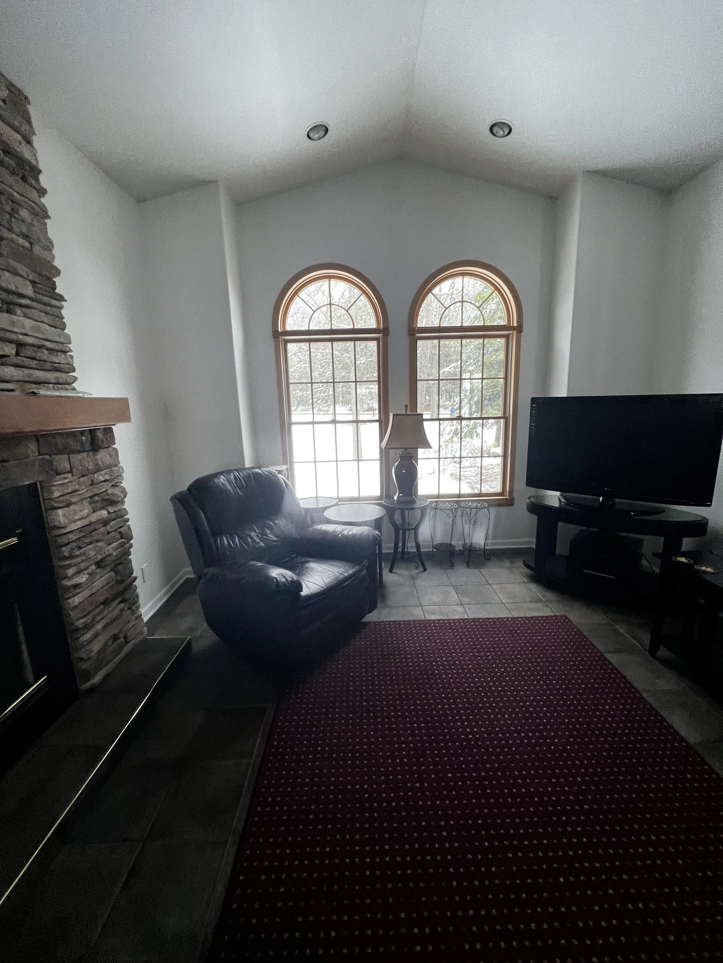 A living room before the transformation with two arched windows, a black leather armchair, a small side table with a lamp, a flat-screen TV on a stand, a red patterned rug, and a stone fireplace on the left.