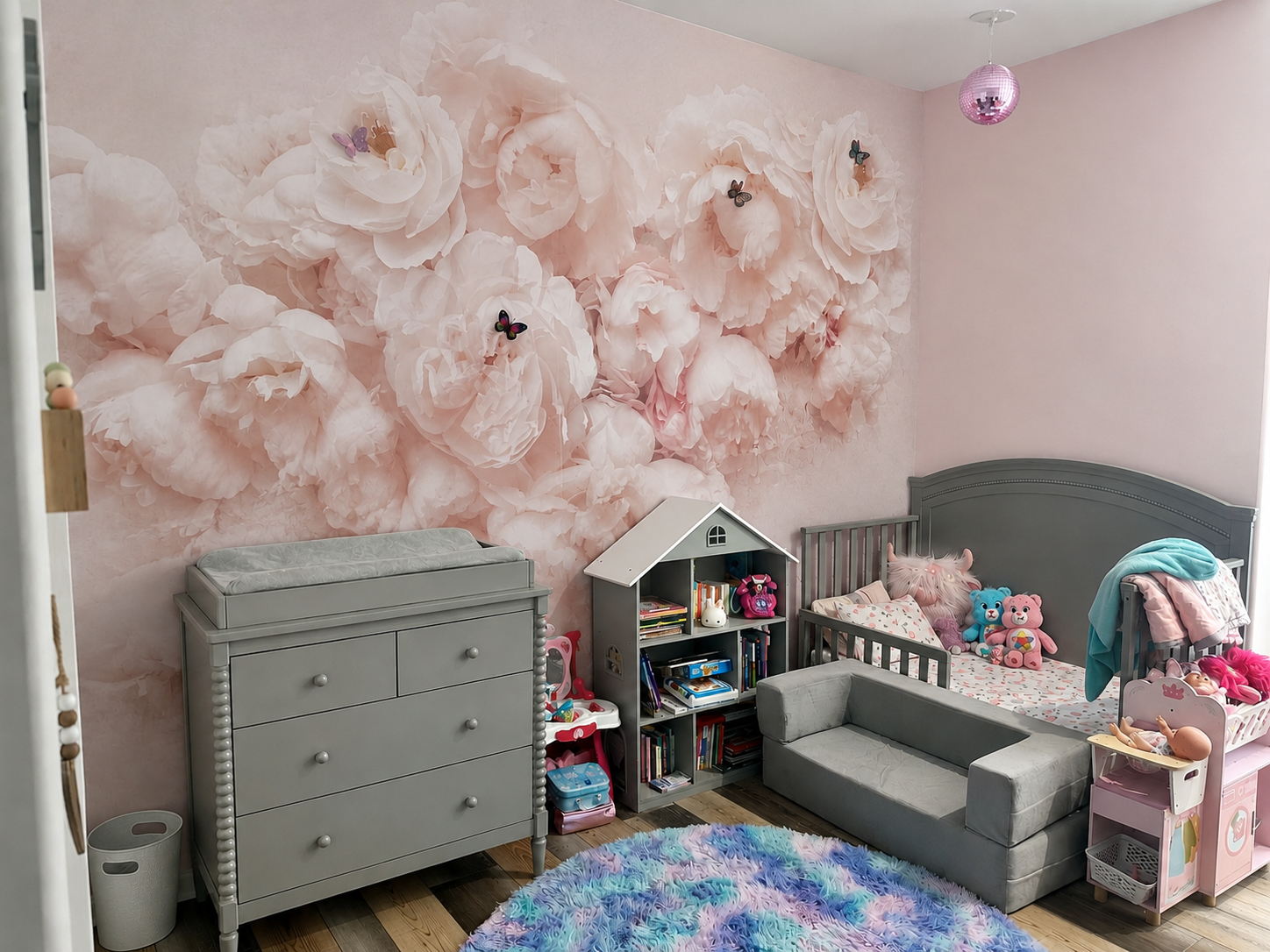 Children's bedroom with pink floral wall mural, toys, books, and a small bed or crib with stuffed animals.