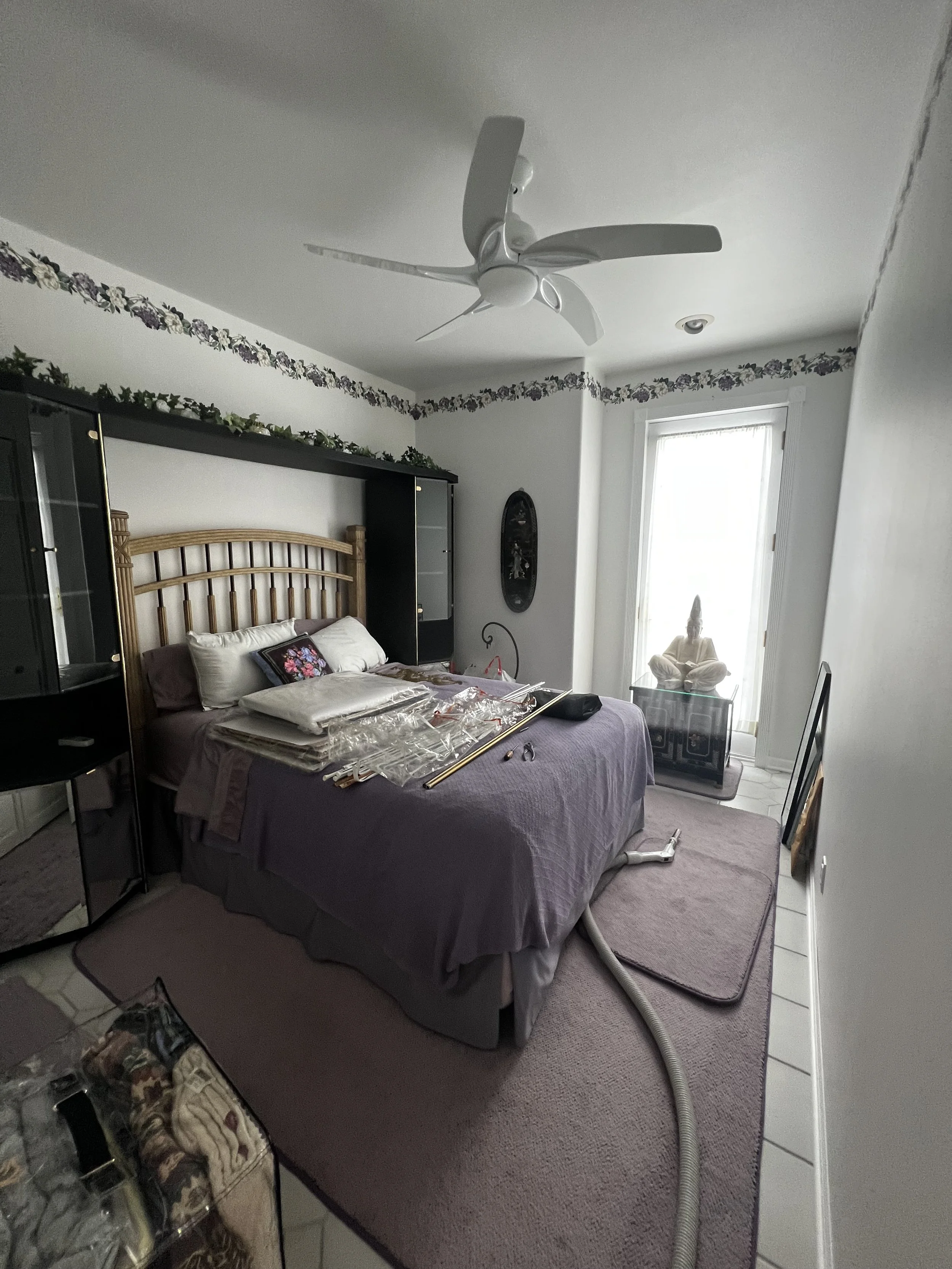 Bedroom before the transformation, featuring a purple bedspread, wall-mounted shelving, a window with sheer curtains, and a simple wall behind the bed before the addition of the princess-themed accent wall.