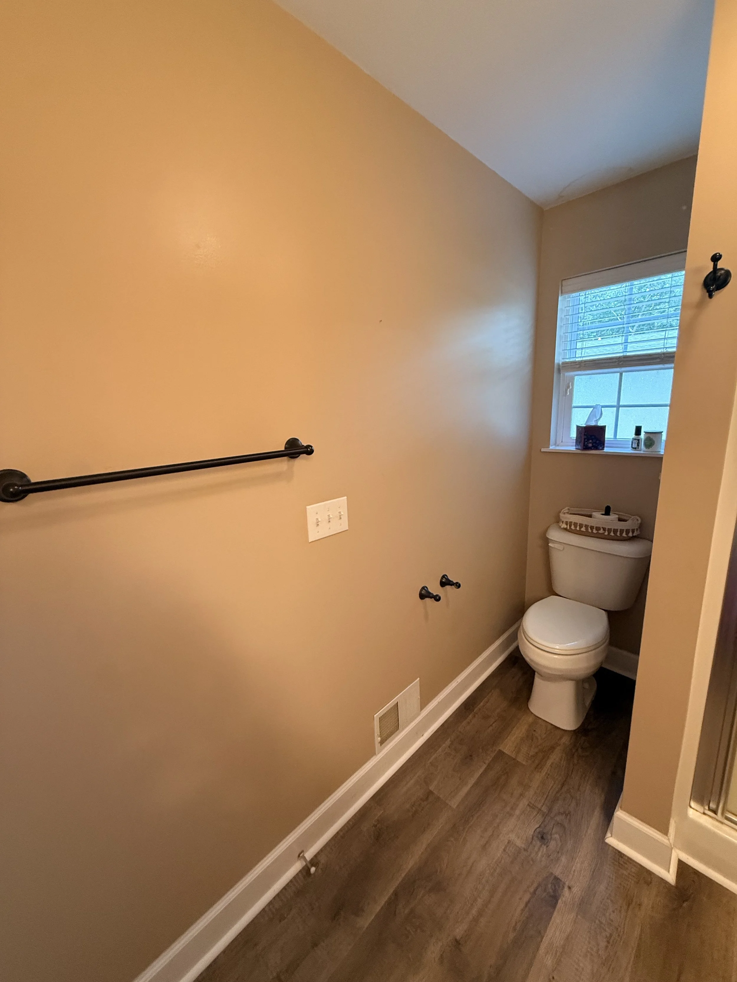 A small bathroom with yellow walls, a window with blinds, a toilet with a tissue box and other items on top, a black handrail on the wall, and hardwood flooring.