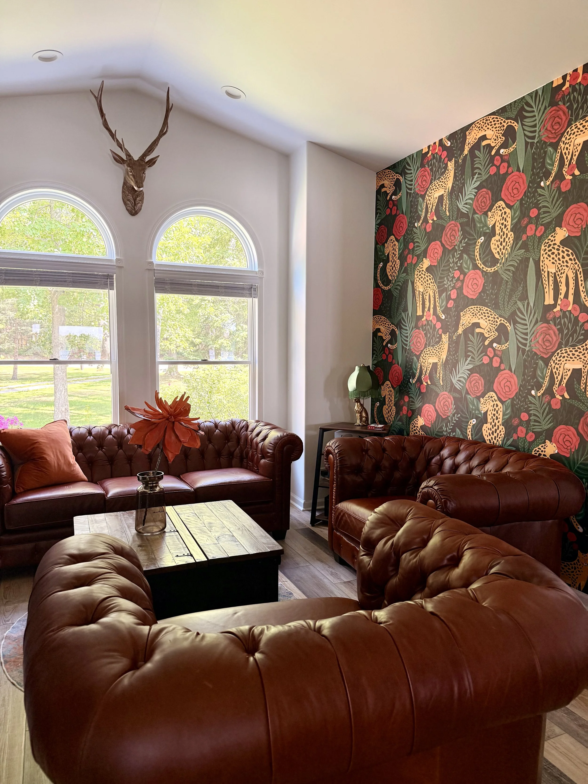 Living room after the transformation, featuring two brown leather sofas and a bold leopard-and-rose wallpaper accent wall, with a wood coffee table, natural light, and warm, inviting details.