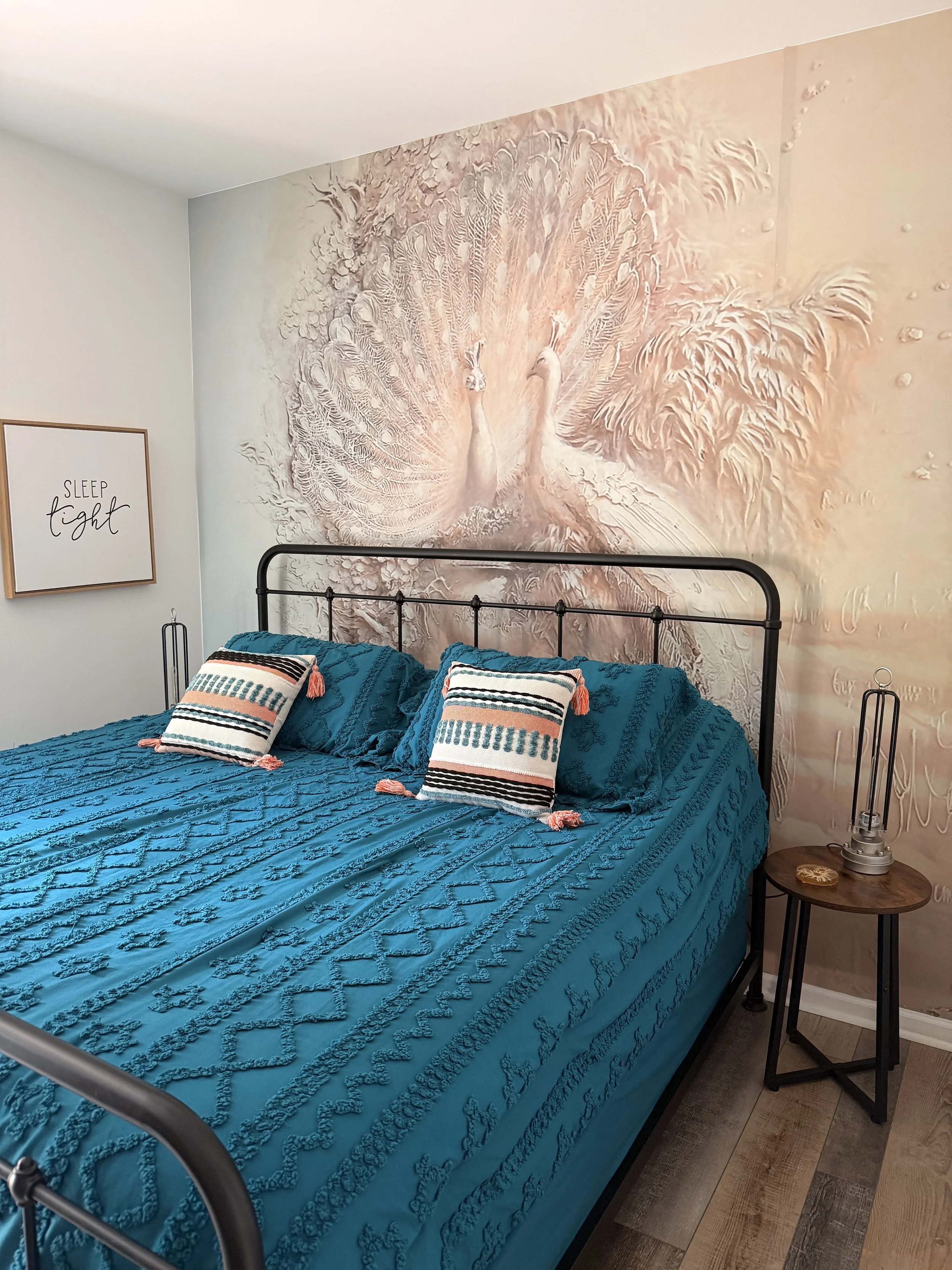Bedroom after the transformation, featuring a colorful turquoise bed, black metal bedframe, and a striking pastel mural of two white peacocks behind the bed, creating a soft and elegant accent wall.