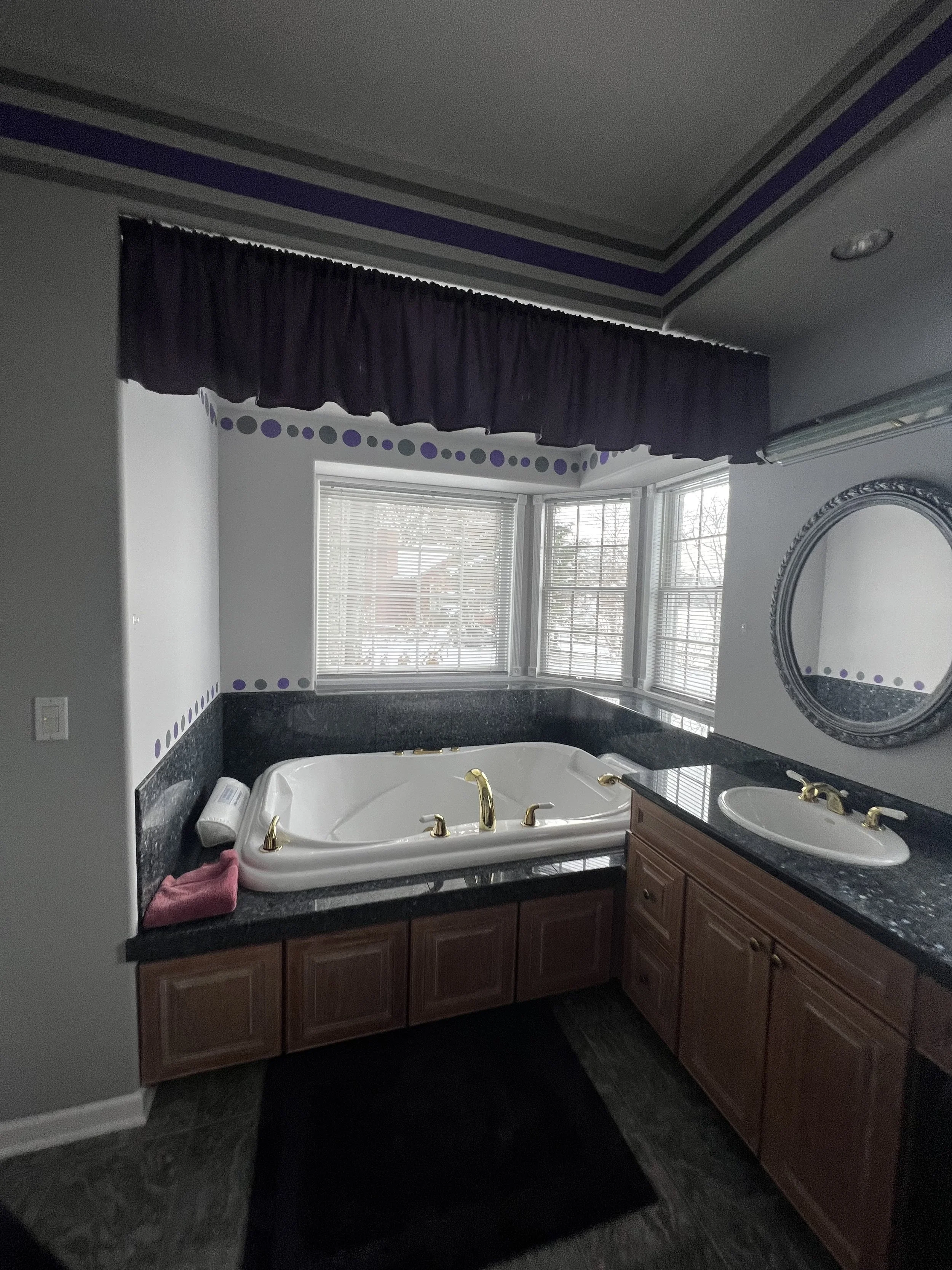 Bathroom before the transformation, featuring a large bathtub with gold fixtures, dark wood cabinetry, a black granite countertop, and a window with blinds, before the addition of the floral accent wall.