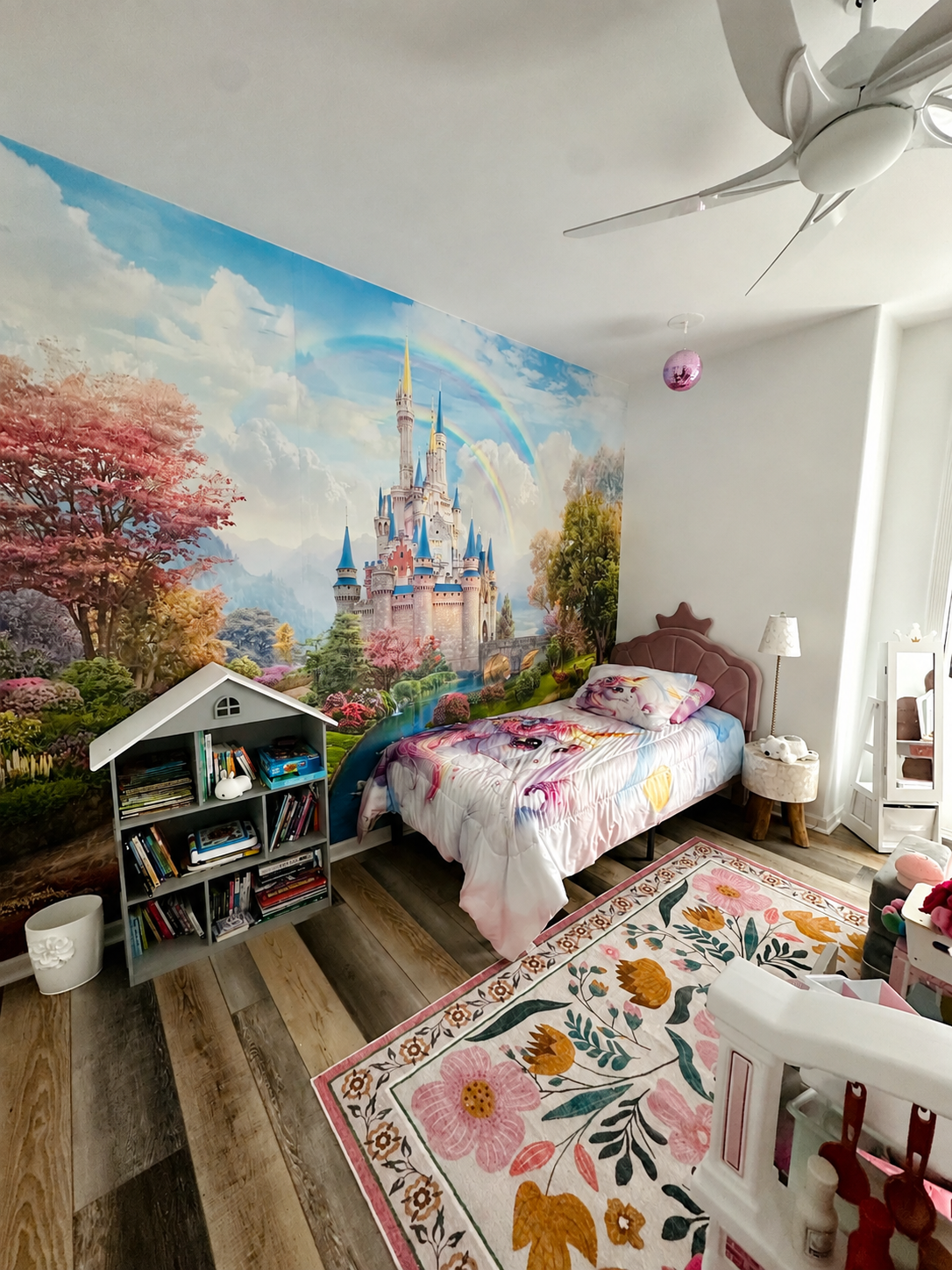A children's bedroom with a colorful fairy tale castle mural on the wall, a twin bed with a unicorn themed bedsheet, a pink headboard, a floral area rug, a small bookshelf, a white stool, and a ceiling fan with a pink ornament hanging.