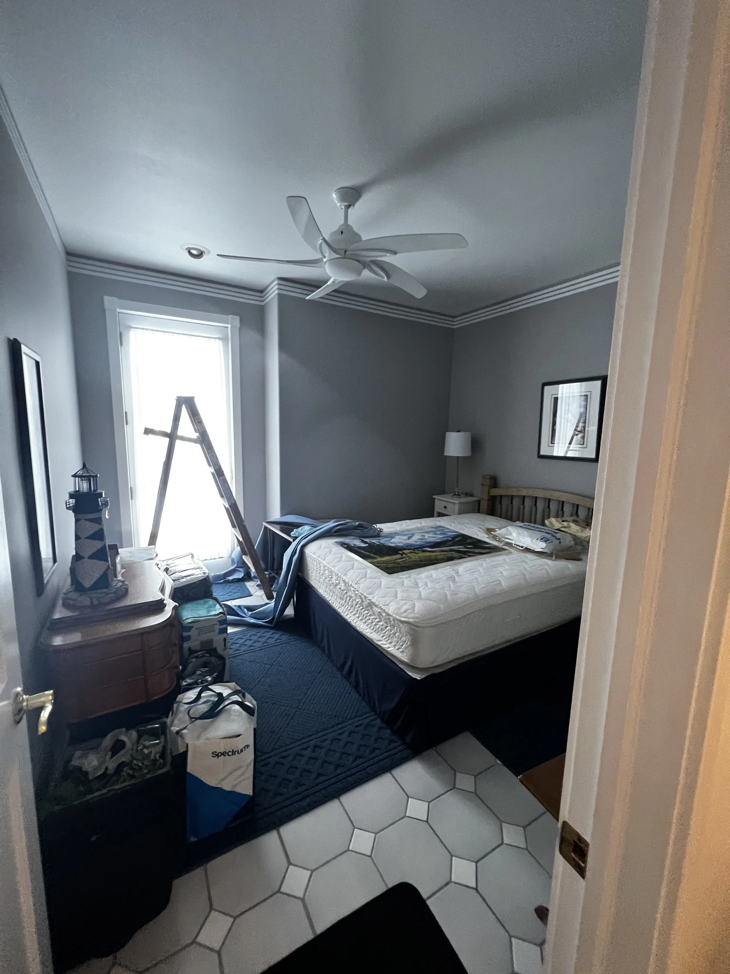 Bedroom before the transformation, with a simple bed, nightstand, lamp, ladder, boxes, and clothing, before the addition of the peacock mural accent wall.