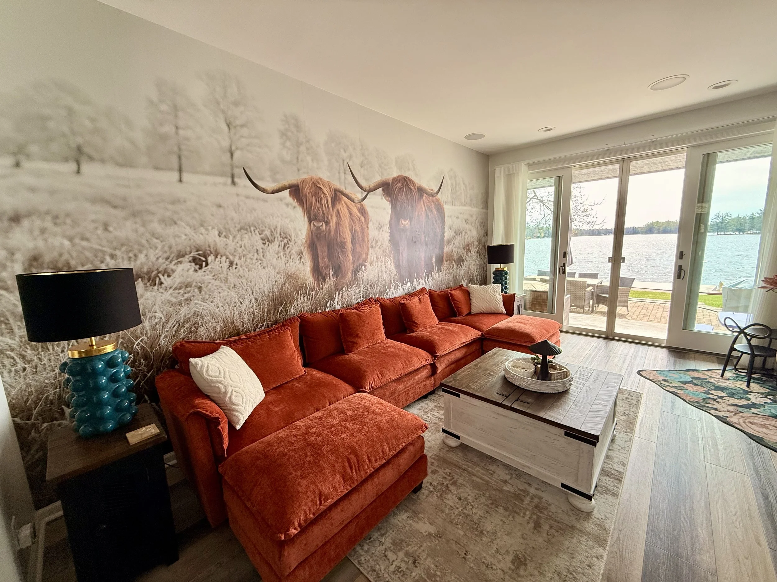 Living room after the transformation, featuring a dramatic Highland cattle mural accent wall, red sofa, black lamps, coffee table, and lake views through sliding glass doors.