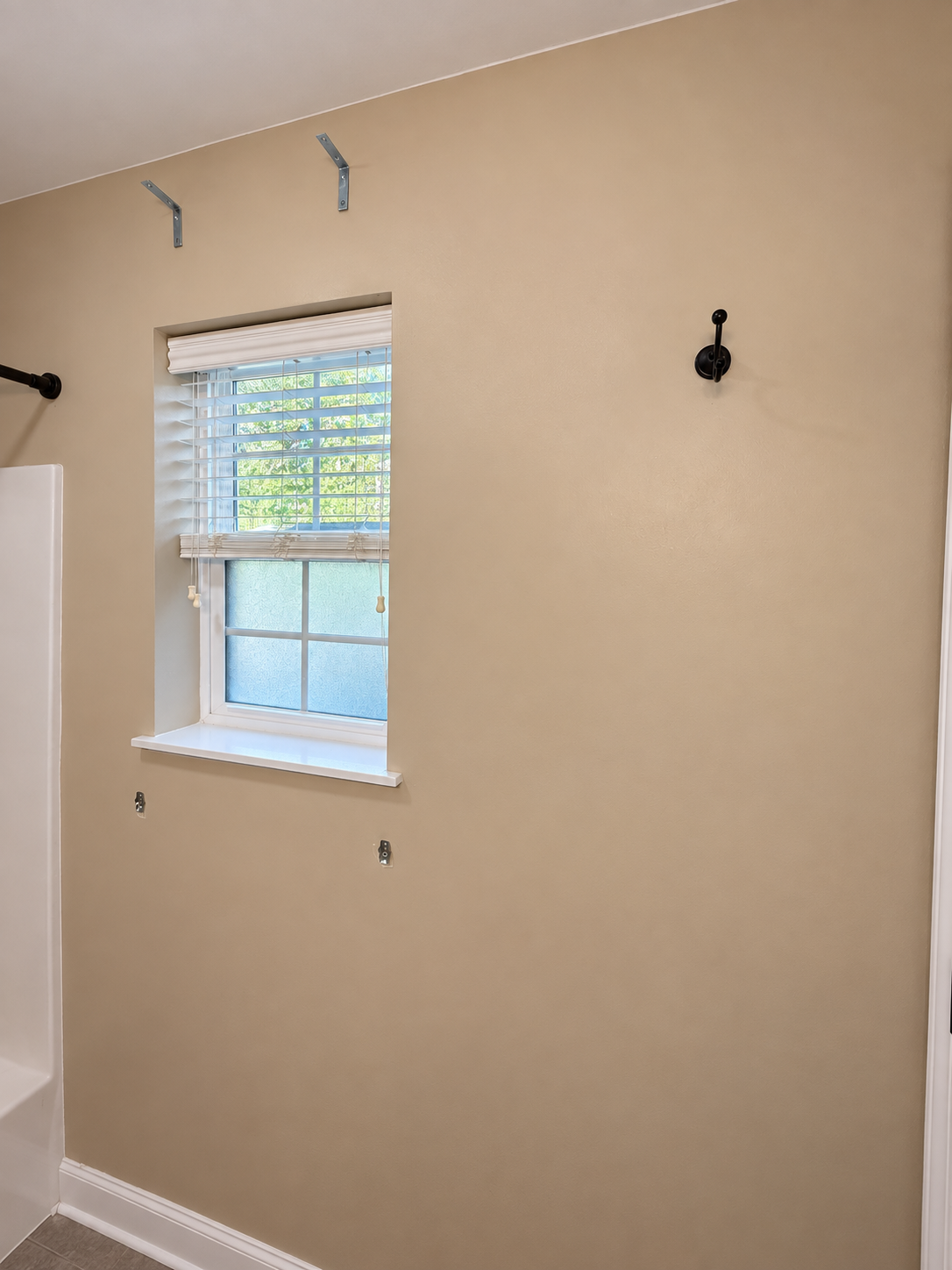 Empty beige wall with a window and black hooks, with metal brackets on the ceiling for hanging items, in a room with beige painted walls.