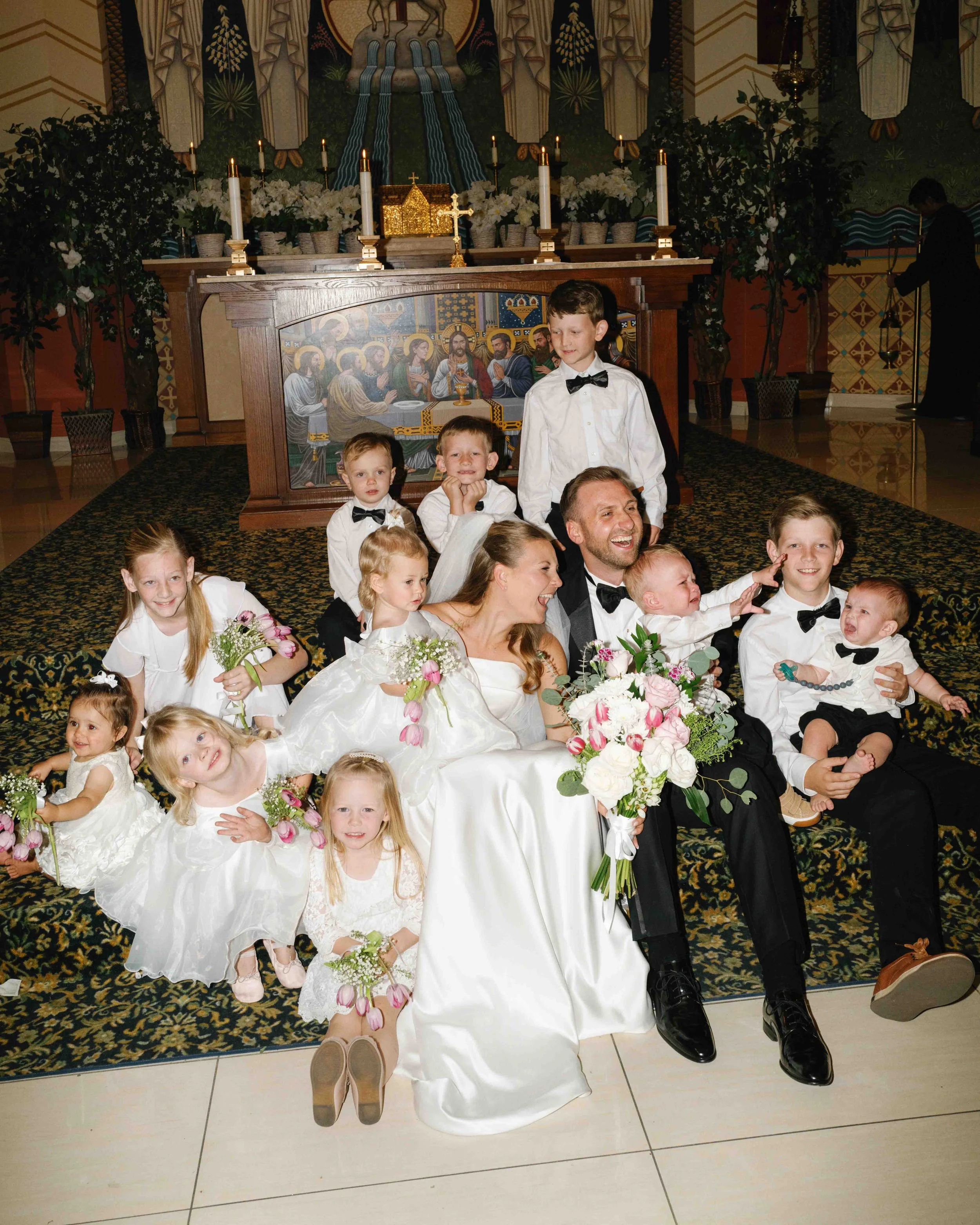 A group of children and a woman dressed in white with a bouquet pose on the floor in front of a church altar decorated with candles, flowers, and religious artwork, during a wedding celebration.