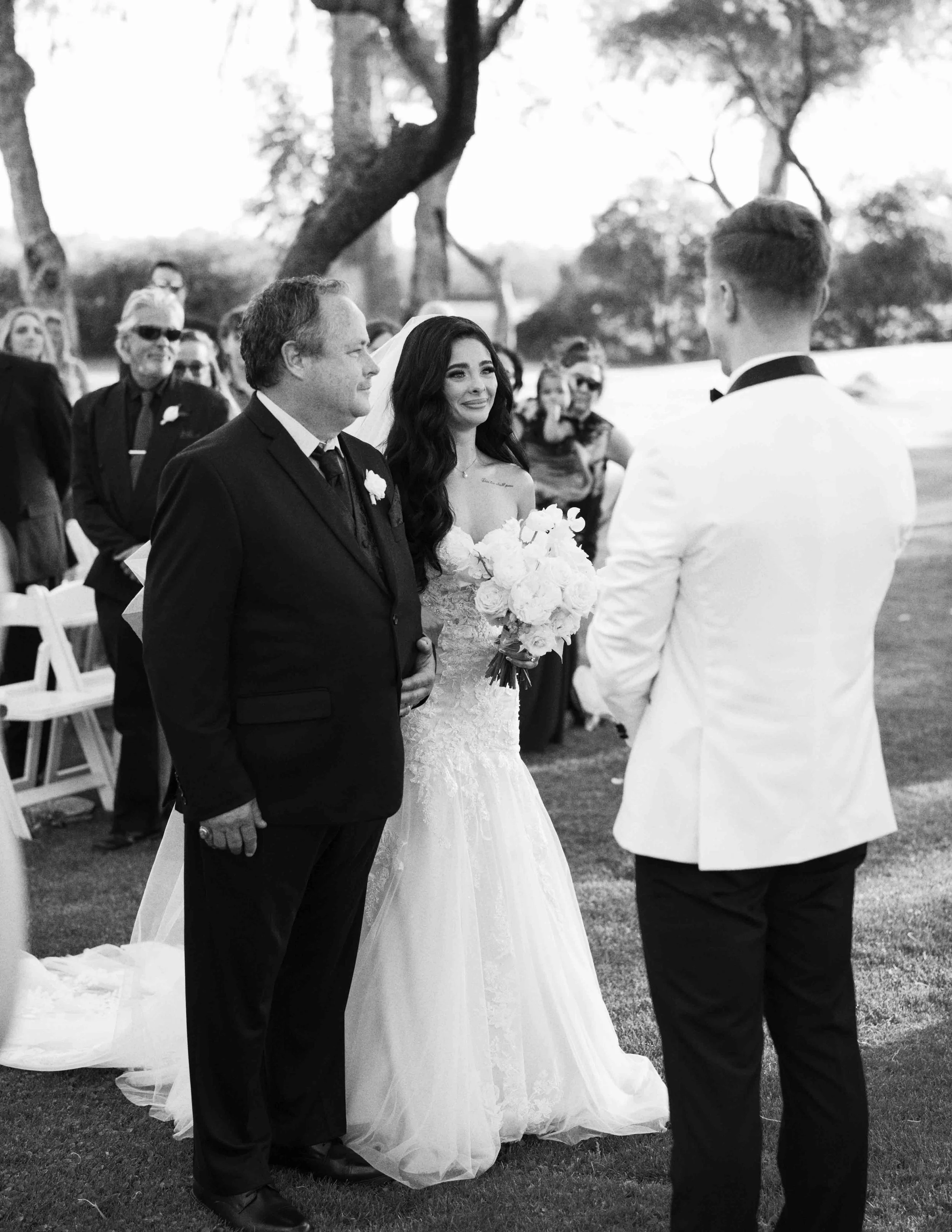 A black and white photo of a wedding ceremony outdoors. The bride, dressed in a wedding gown and holding a bouquet of flowers, faces the groom, dressed in a white tuxedo. An older man in a black suit stands nearby. Guests are seated or standing in th
