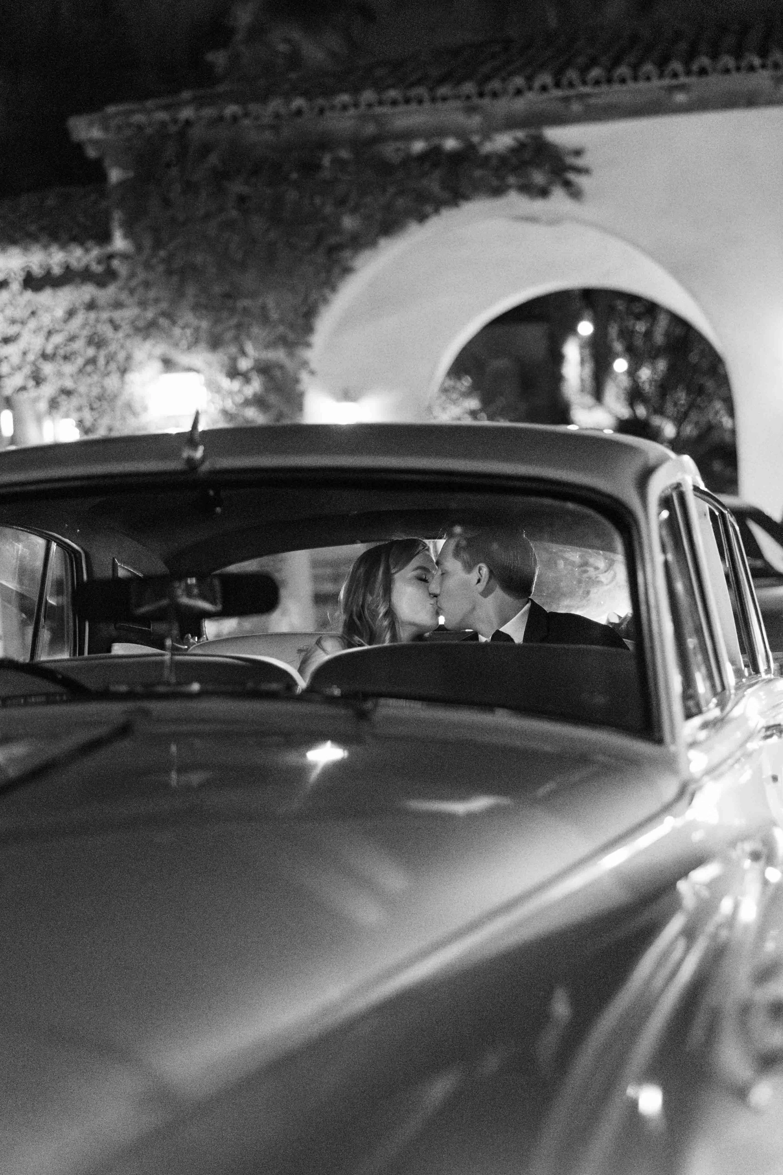 A black and white photo of a vintage car with a couple inside, about to kiss, in front of an arched structure at night.