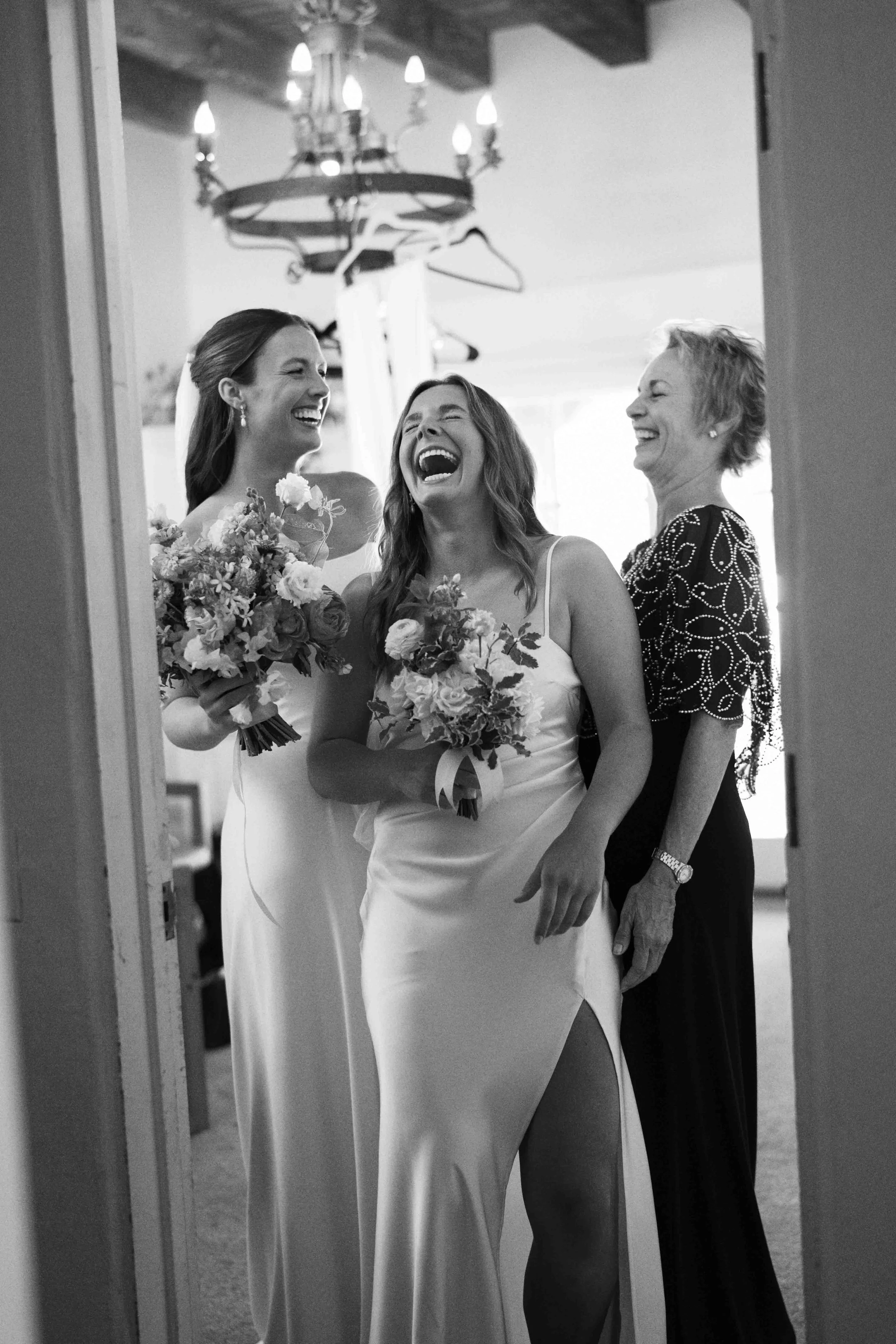 Three women, including a bride in a wedding dress, laughing and smiling as they look at each other in a room with a chandelier overhead.