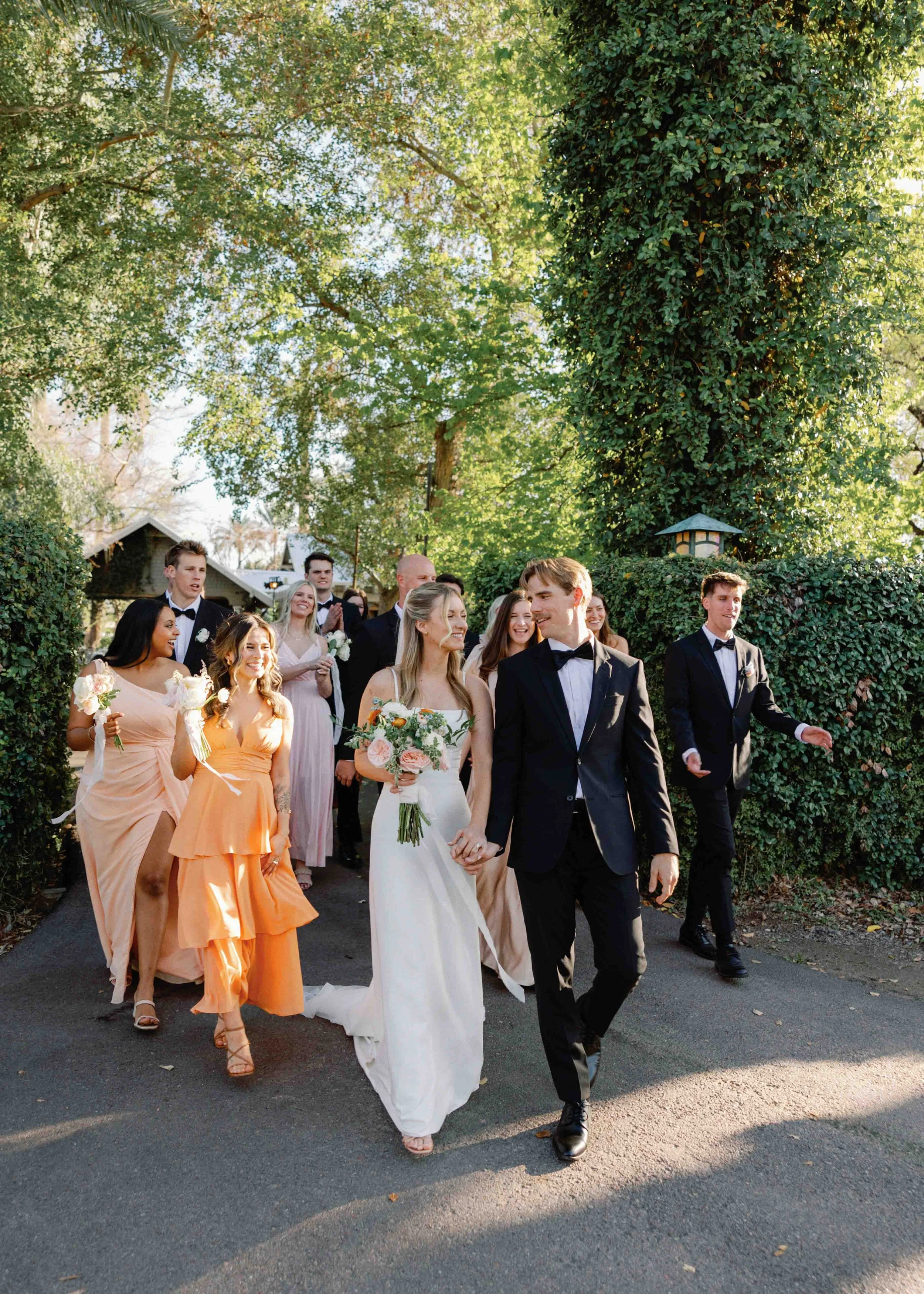 A wedding party walking outdoors on a sunny day, with the bride in a white dress holding a bouquet, and the groom in a tuxedo. Bridesmaids and groomsmen are dressed in pastel and black attire, surrounded by trees and greenery.