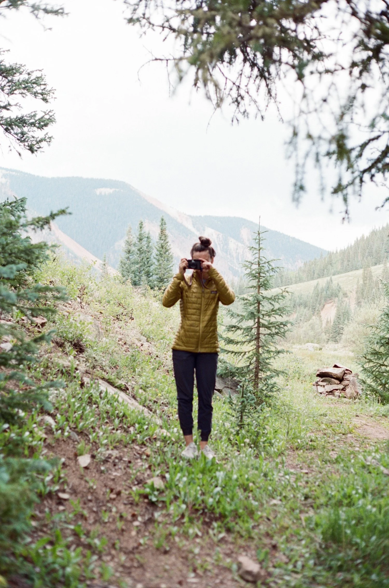A person standing on a trail in a forest, taking a photo with a camera. They are wearing a green puffer jacket, black pants, and white shoes, with mountains and trees in the background.