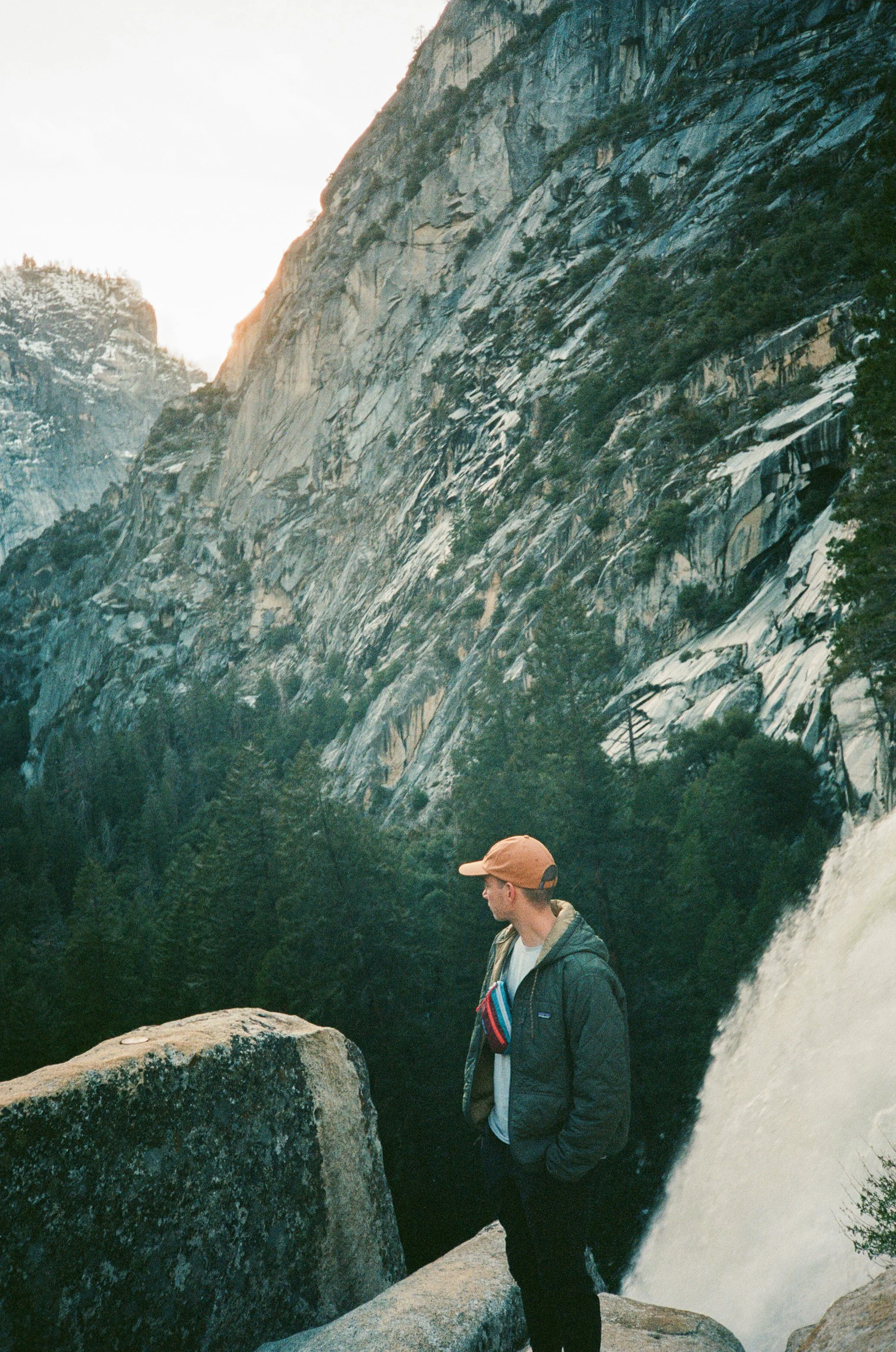 A person wearing a baseball cap, glasses, a jacket, and backpack standing near a large boulder in front of a waterfall with rugged mountain cliffs in the background.