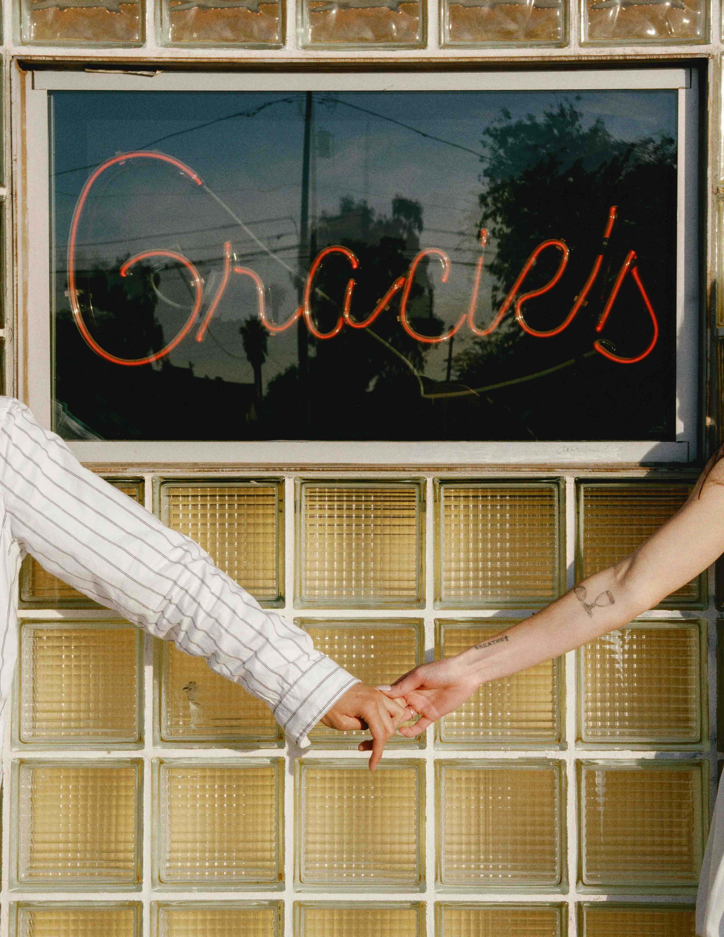 Two people holding hands in front of a glass block wall with a neon sign reading 'accepts' in cursive script. One person's arm has tattoos, including the word 'BREATHE.'