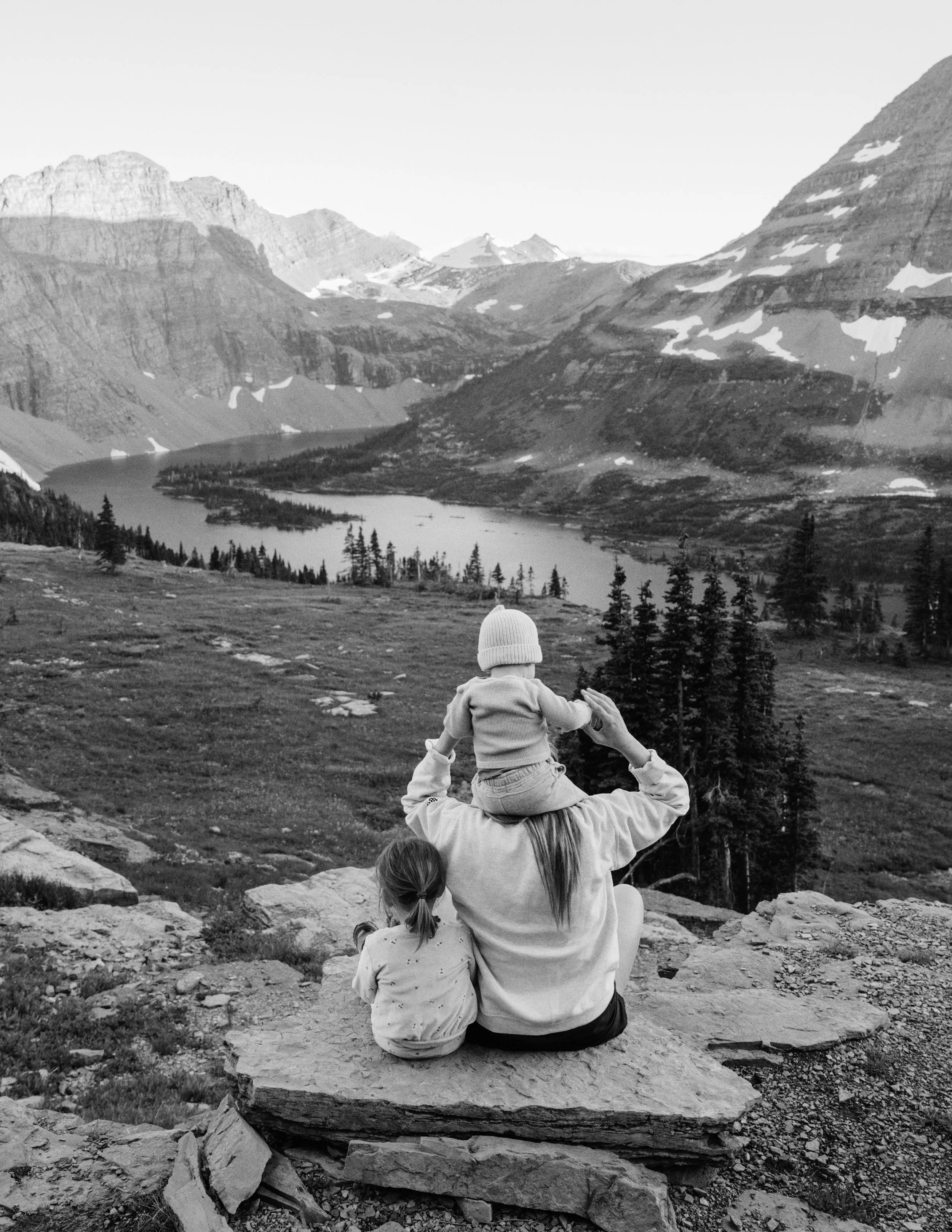 A woman sitting on a rocky ledge with her child on her shoulders, overlooking a lake surrounded by mountains with patches of snow, in black and white.