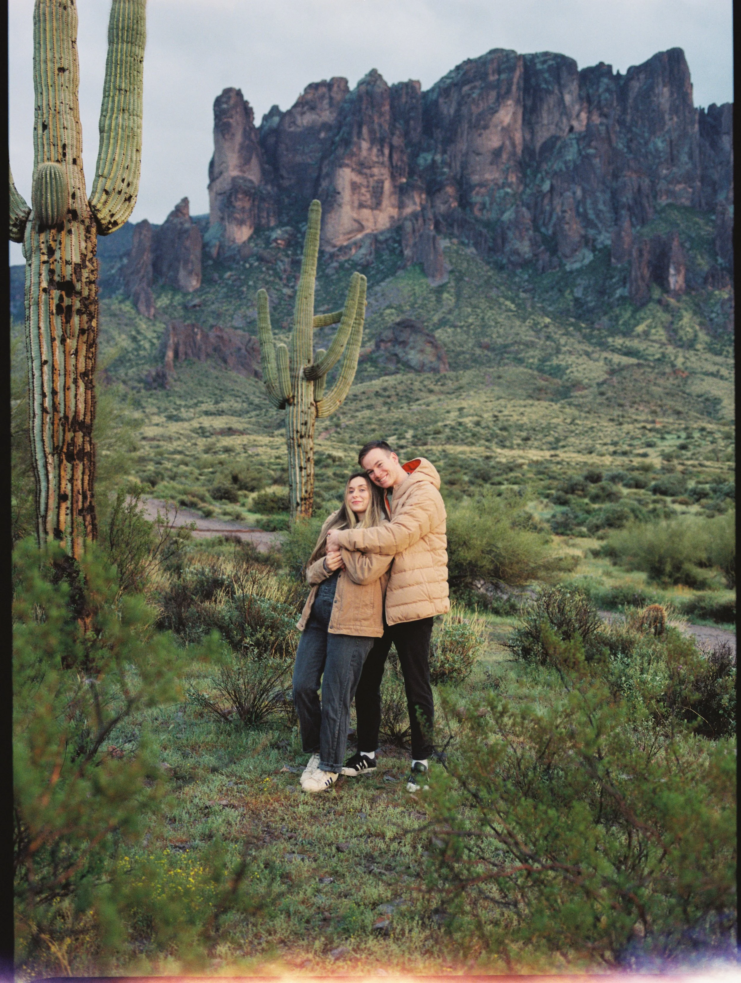 A young couple hugging in a desert landscape with large cacti and rocky mountains in the background.