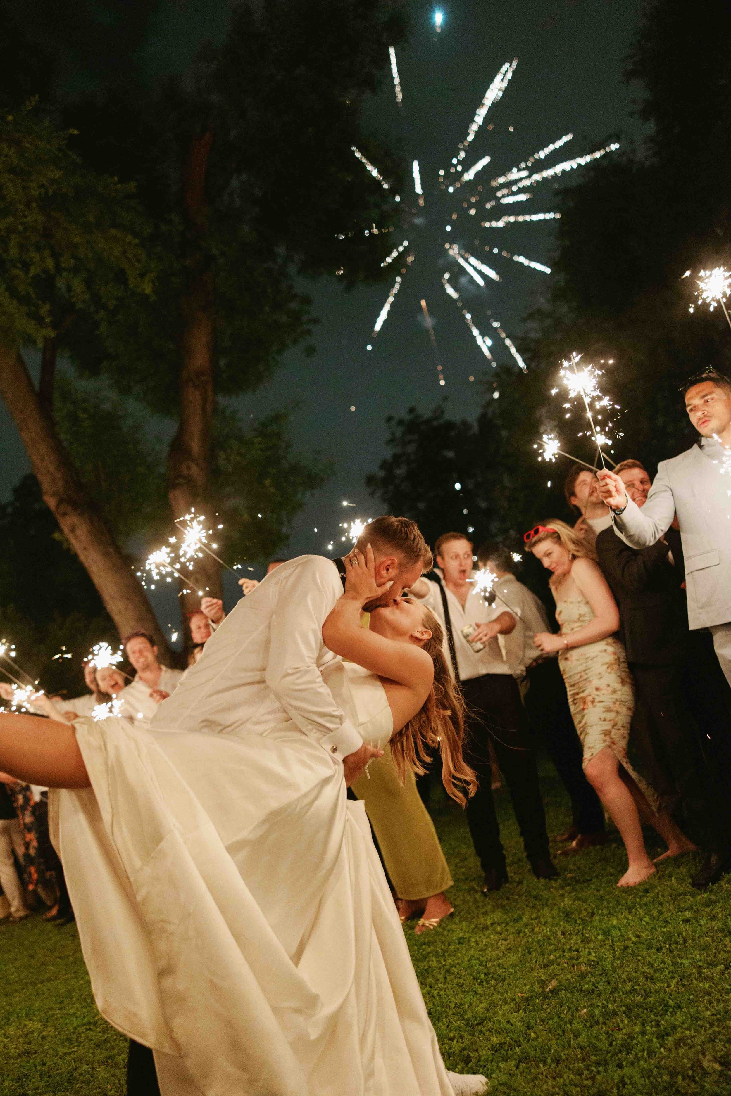 A man and woman in wedding attire sharing a kiss during a celebration, surrounded by guests holding sparklers at night with fireworks in the sky.