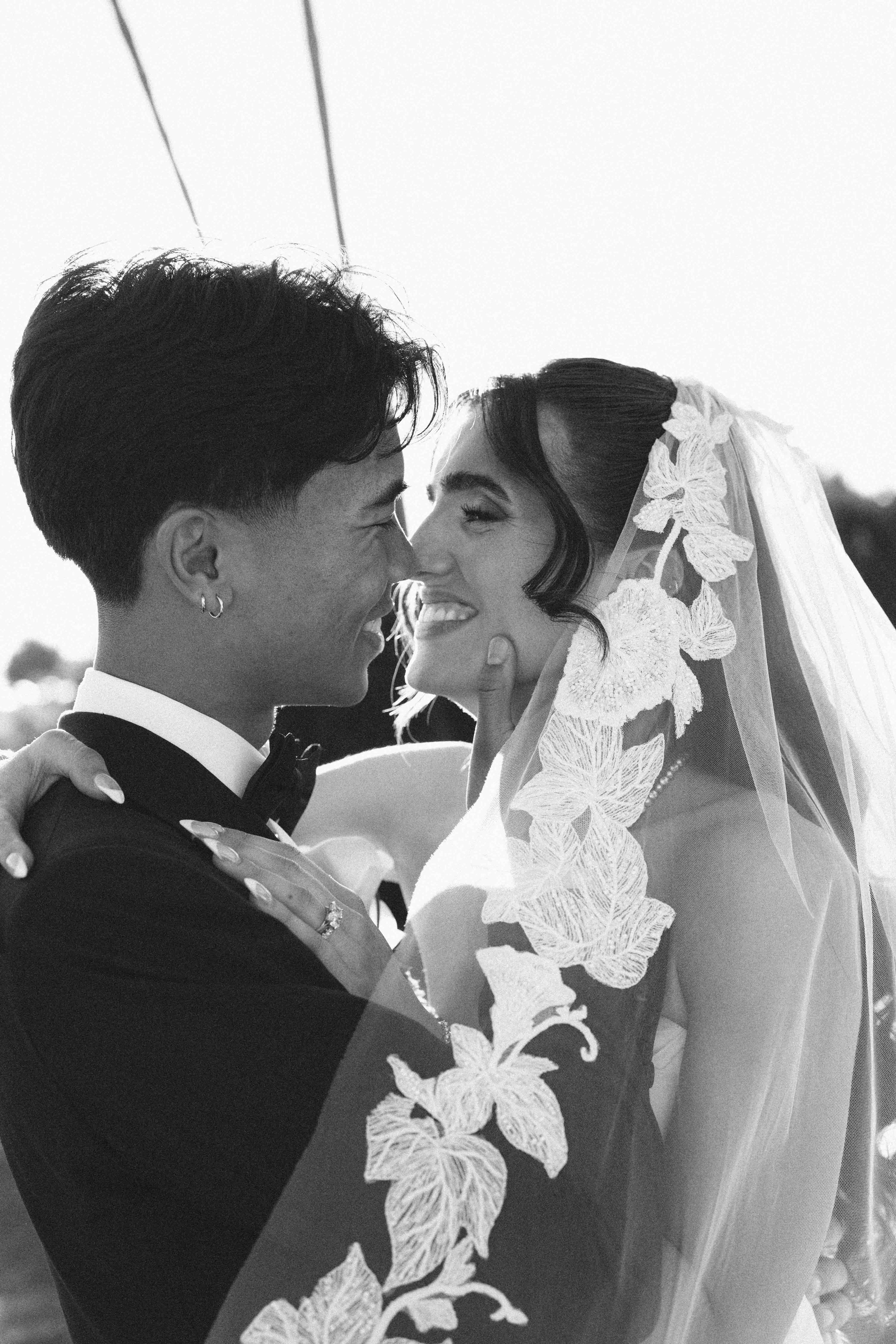 Black and white photo of a bride and groom smiling closely, with the bride wearing a lace veil and flower details, and the groom in a tuxedo with a bow tie.