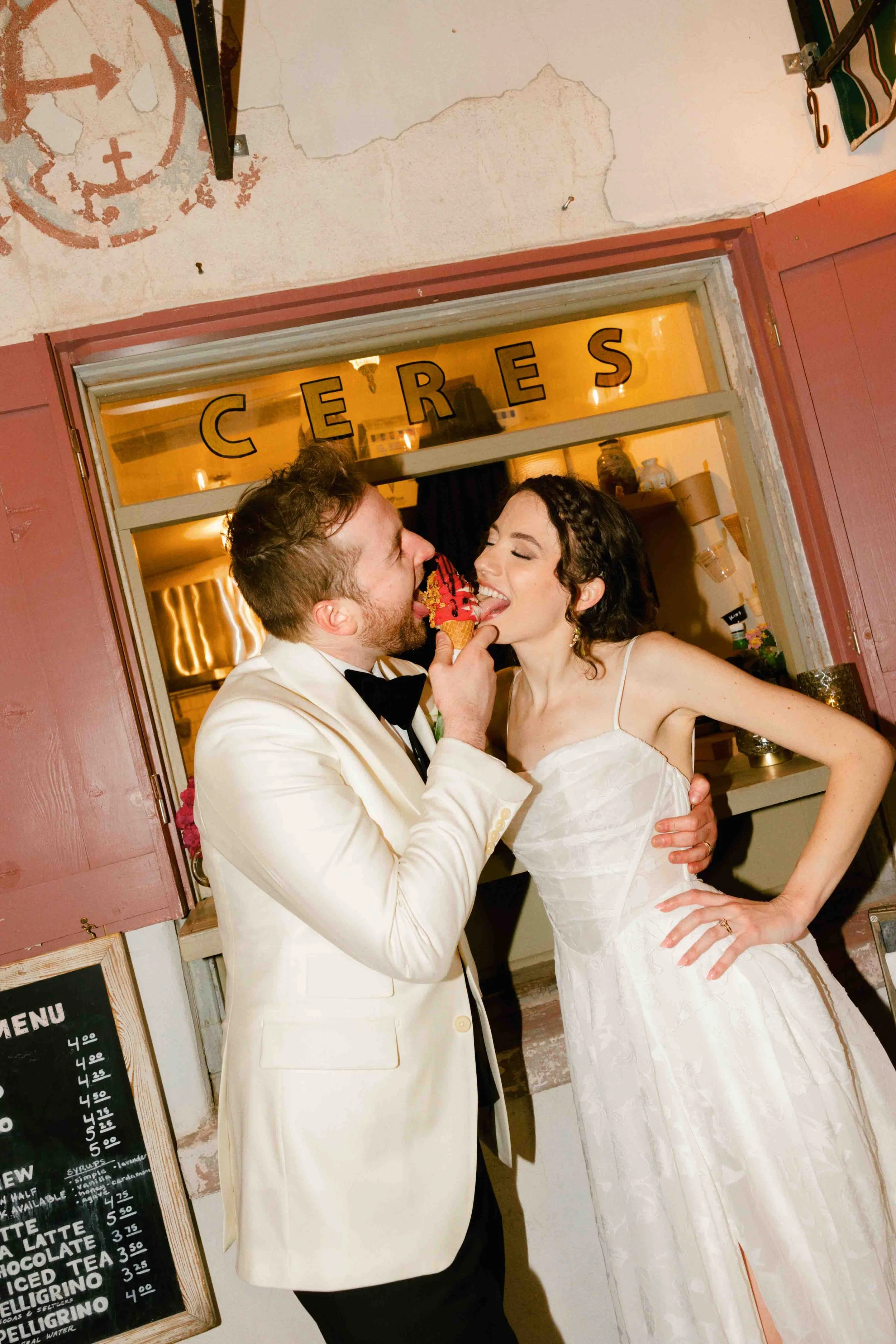 A man in a white tuxedo feeding a woman in a wedding dress ice cream in a cafe or restaurant.