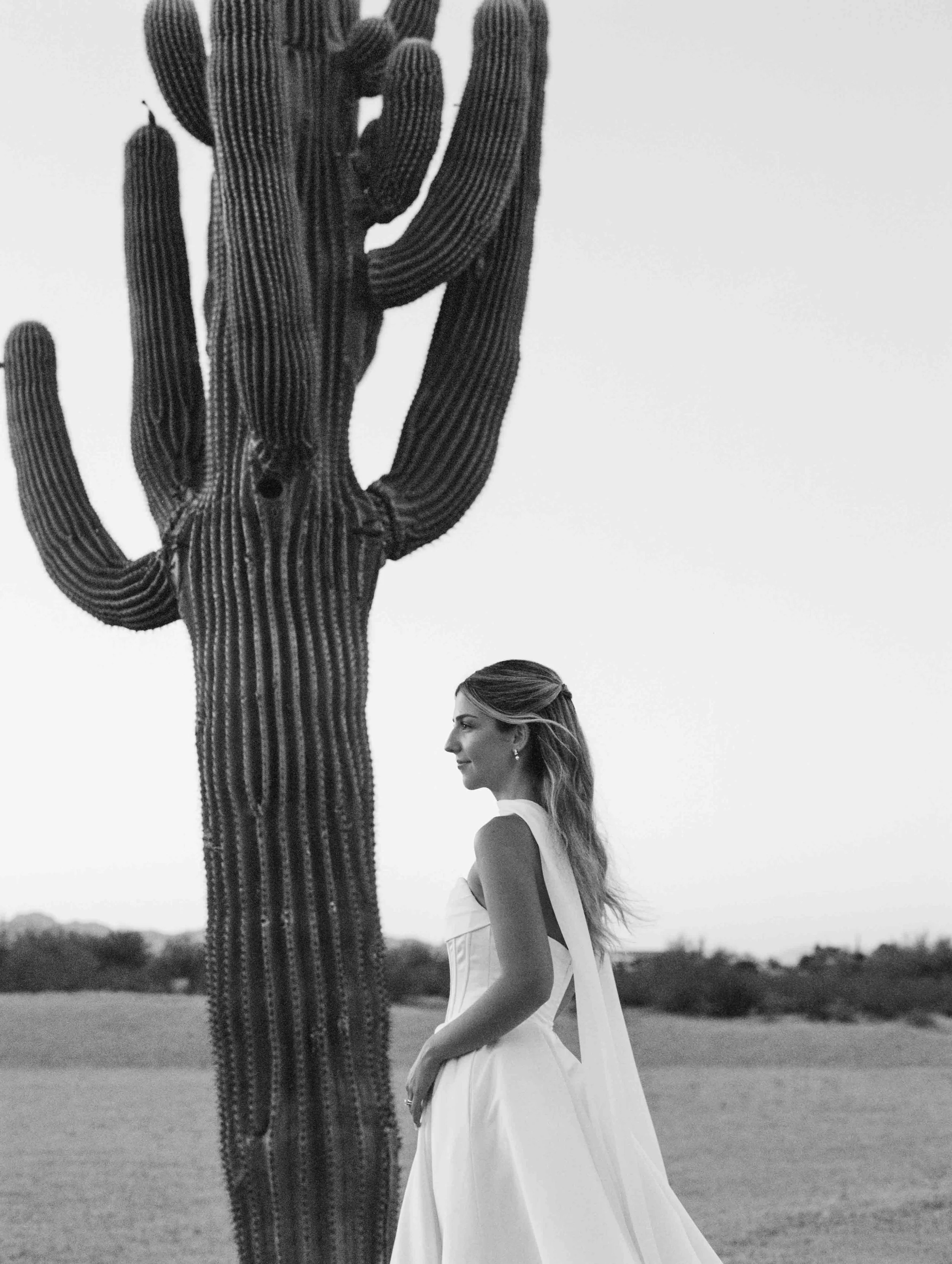 A woman in a white dress standing outdoors and looking to the right, with a large cactus in the background.