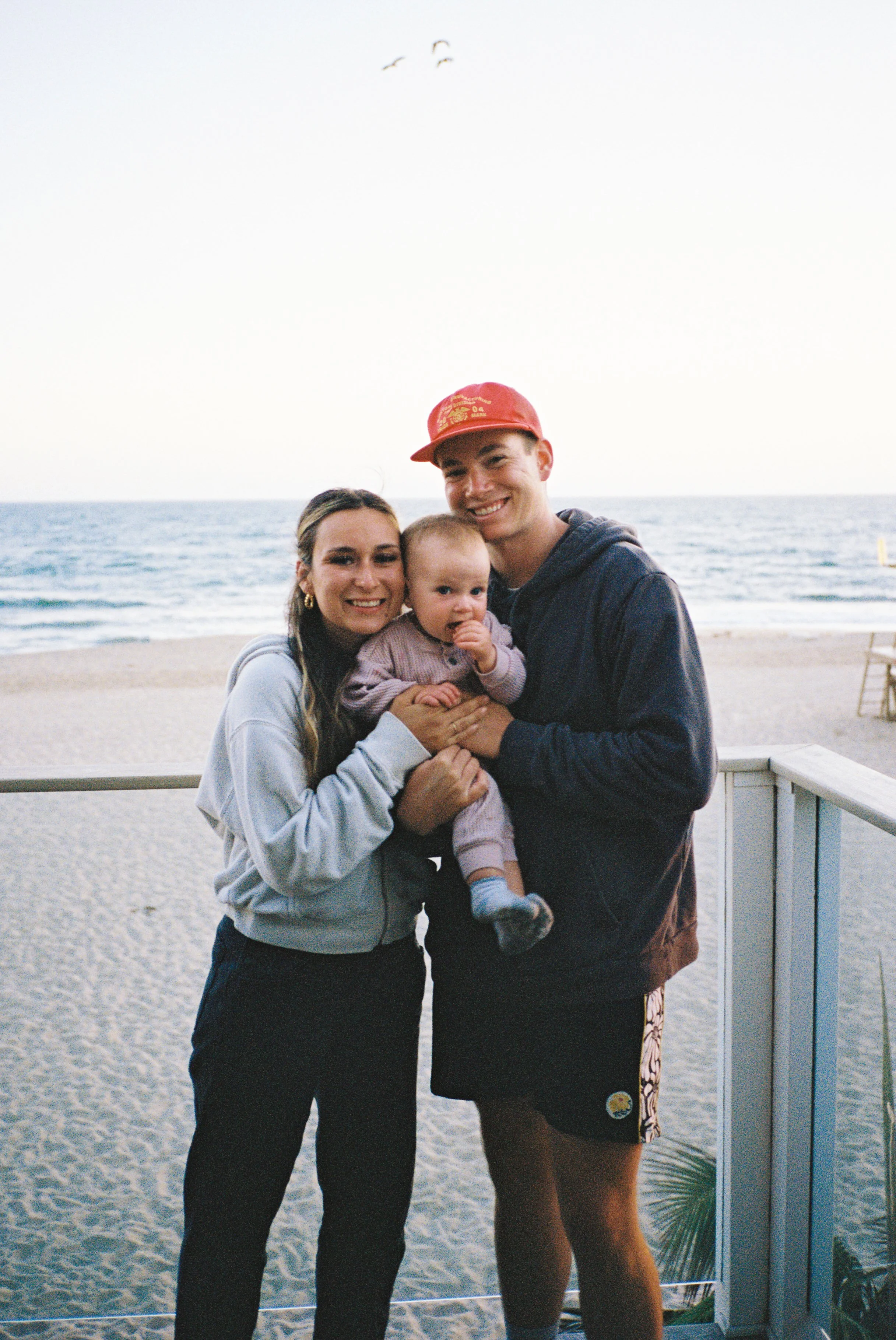 A smiling family of three at the beach, with an ocean and seagulls flying in the sky in the background. The mother, father, and young child are posing together, with the father wearing a red hat and the mother wearing earrings.