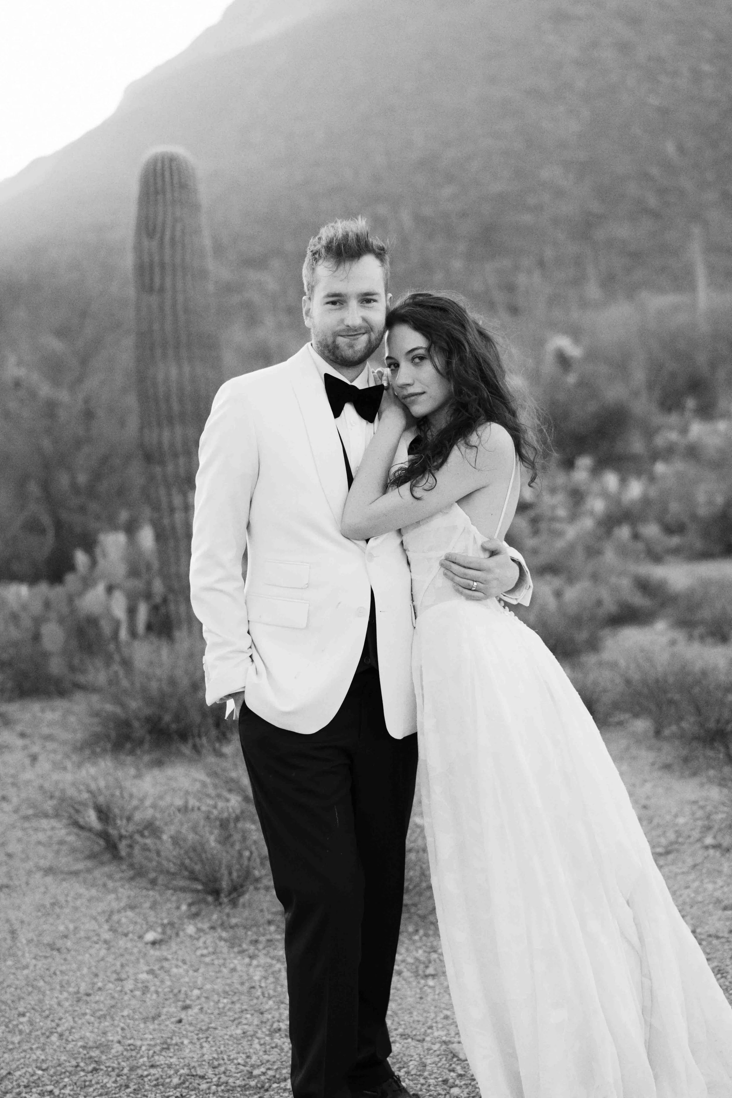 A black and white photo of a couple dressed in formal wedding attire, standing outdoors with desert plants and mountains in the background.