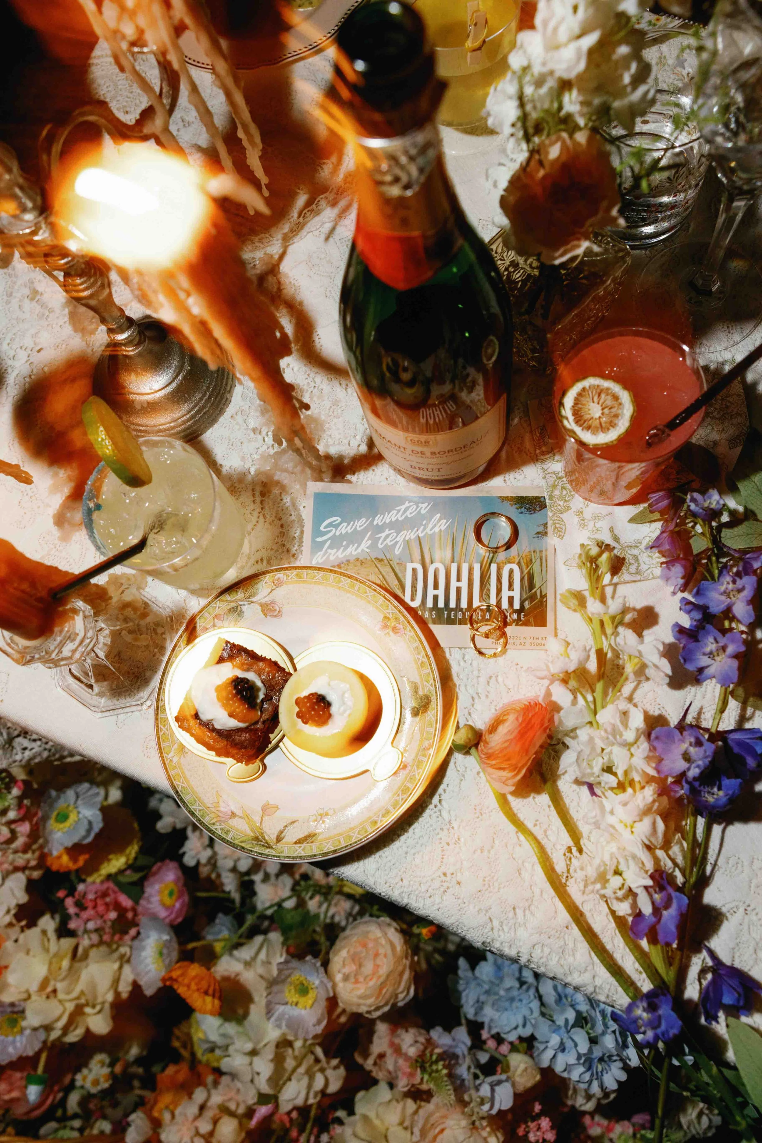 A table set with various drinks, desserts, and surrounded by an abundance of colorful flowers. The drinks include a lemon garnish cocktail, a strawberry cocktail with lemon slice, and a dark cocktail with a lemon slice. There are also small desserts 