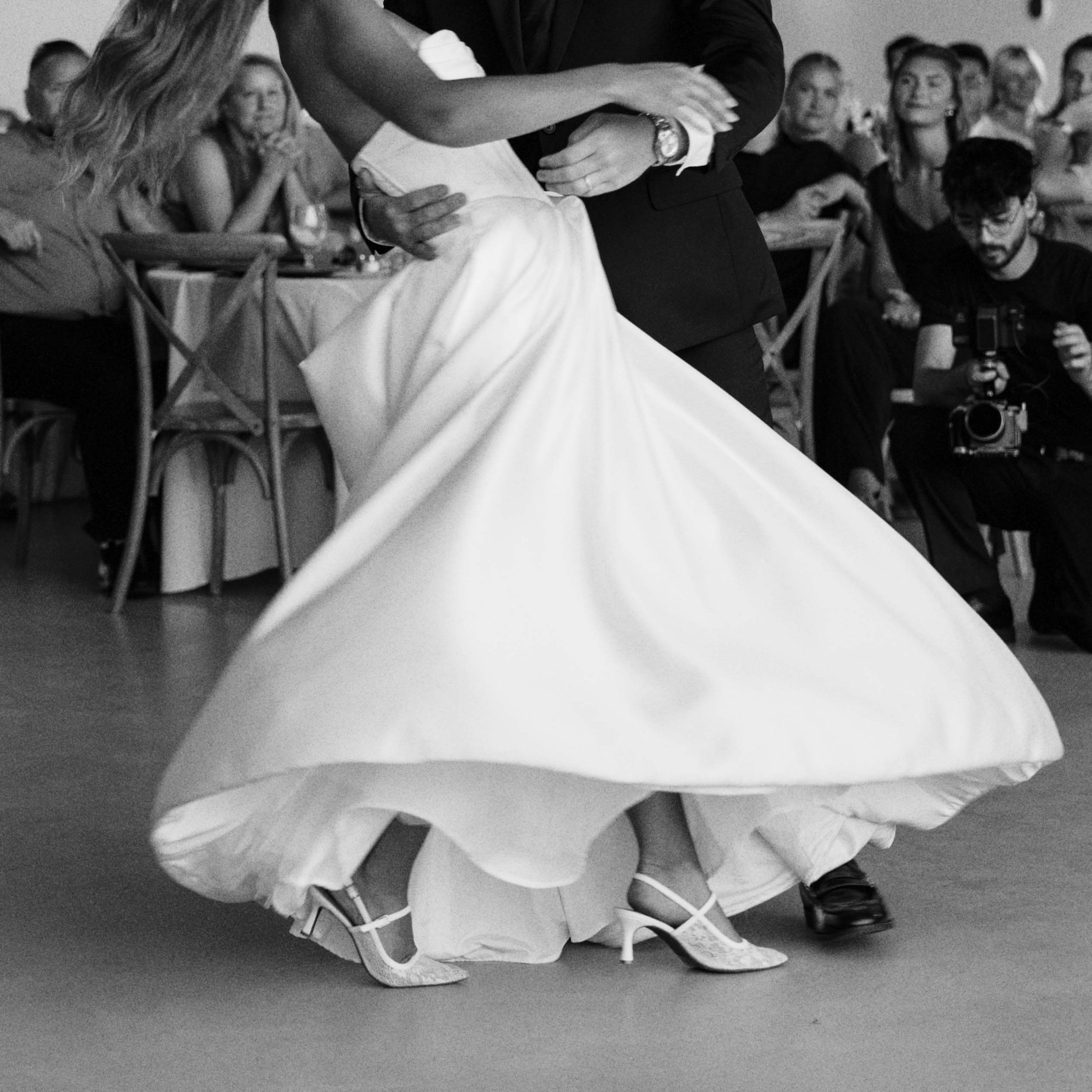 A couple dancing close together at a formal event, with guests seated at tables in the background. The woman is wearing a long gown, and the man is dressed in a suit. A photographer is capturing the moment.
