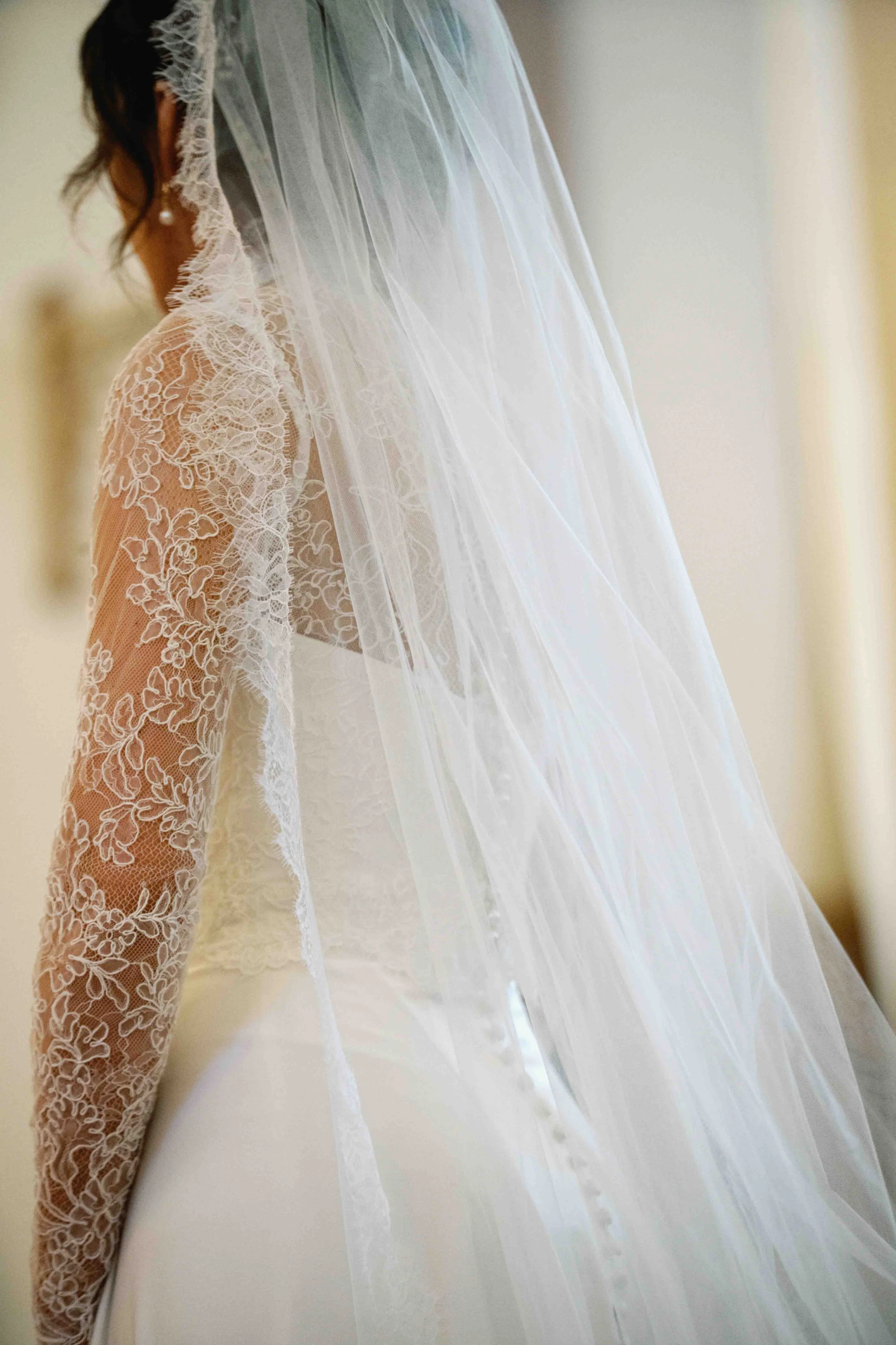 A woman wearing a wedding dress with lace long sleeves and a veil standing near a beige wall.