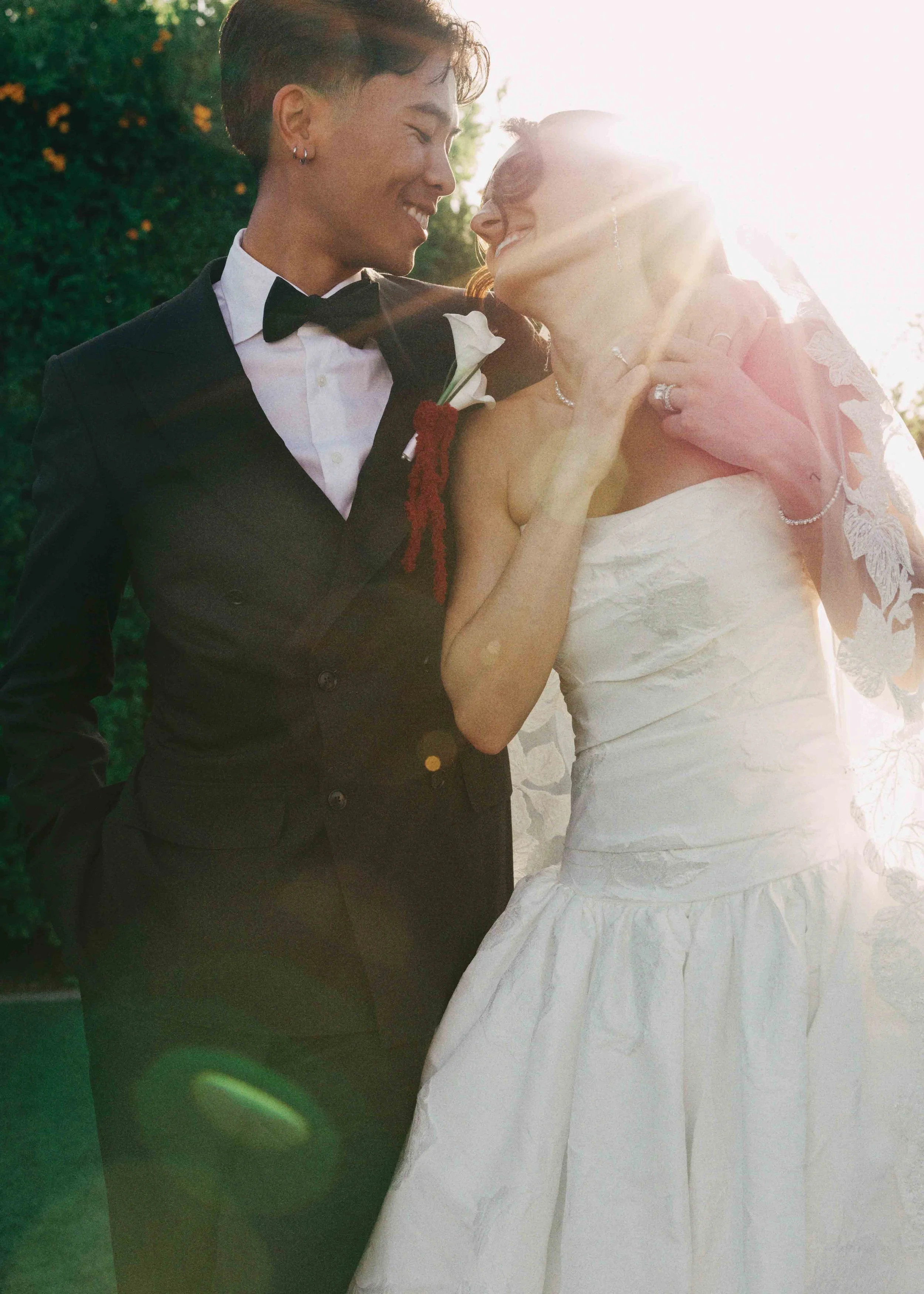 A bride and groom share a tender moment, smiling at each other, outside during sunset, with the bride wearing a white wedding gown and the groom in a tuxedo.