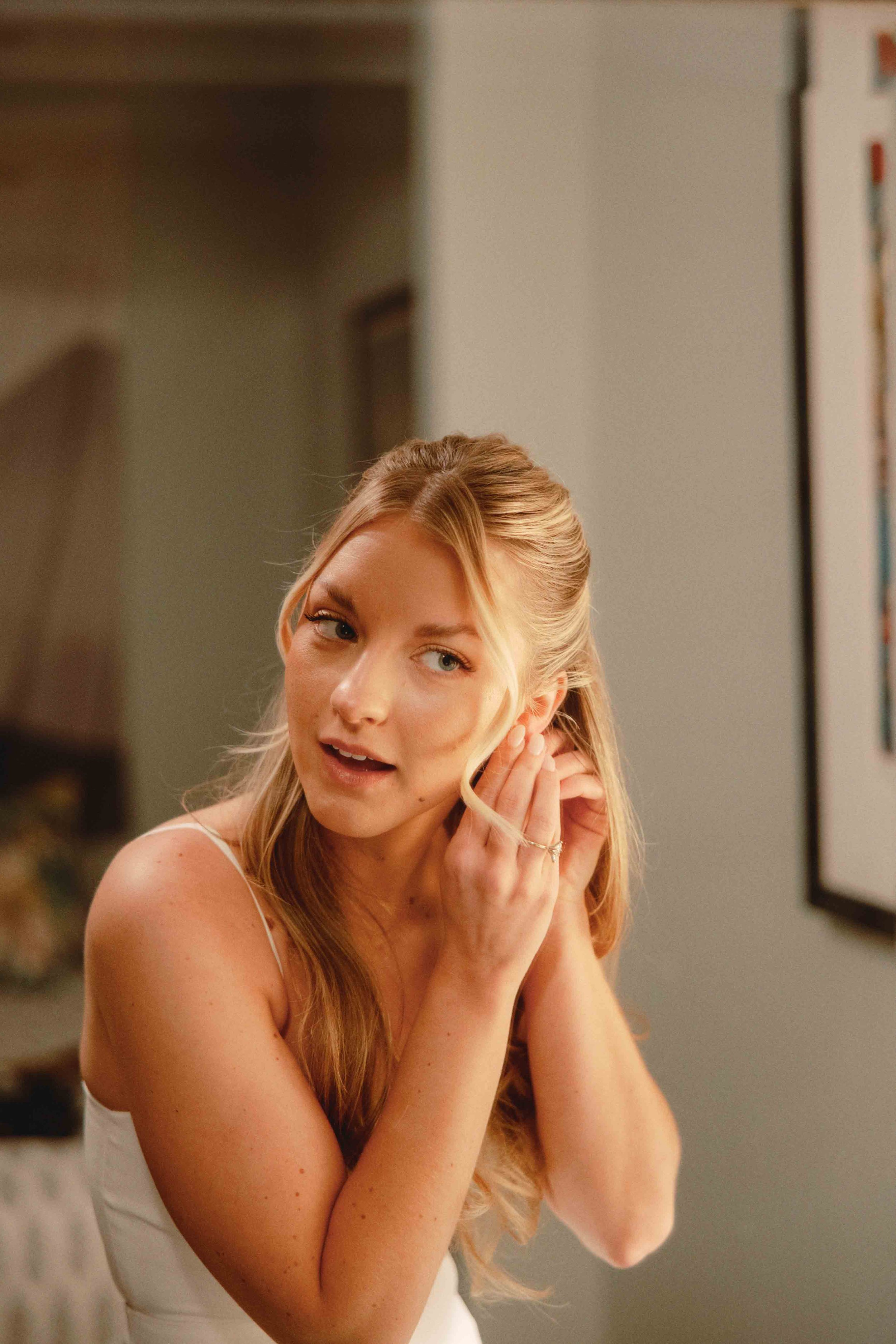A blonde woman wearing a white sleeveless top adjusting her hair with her right hand, smiling and looking slightly upward in a warmly lit room.
