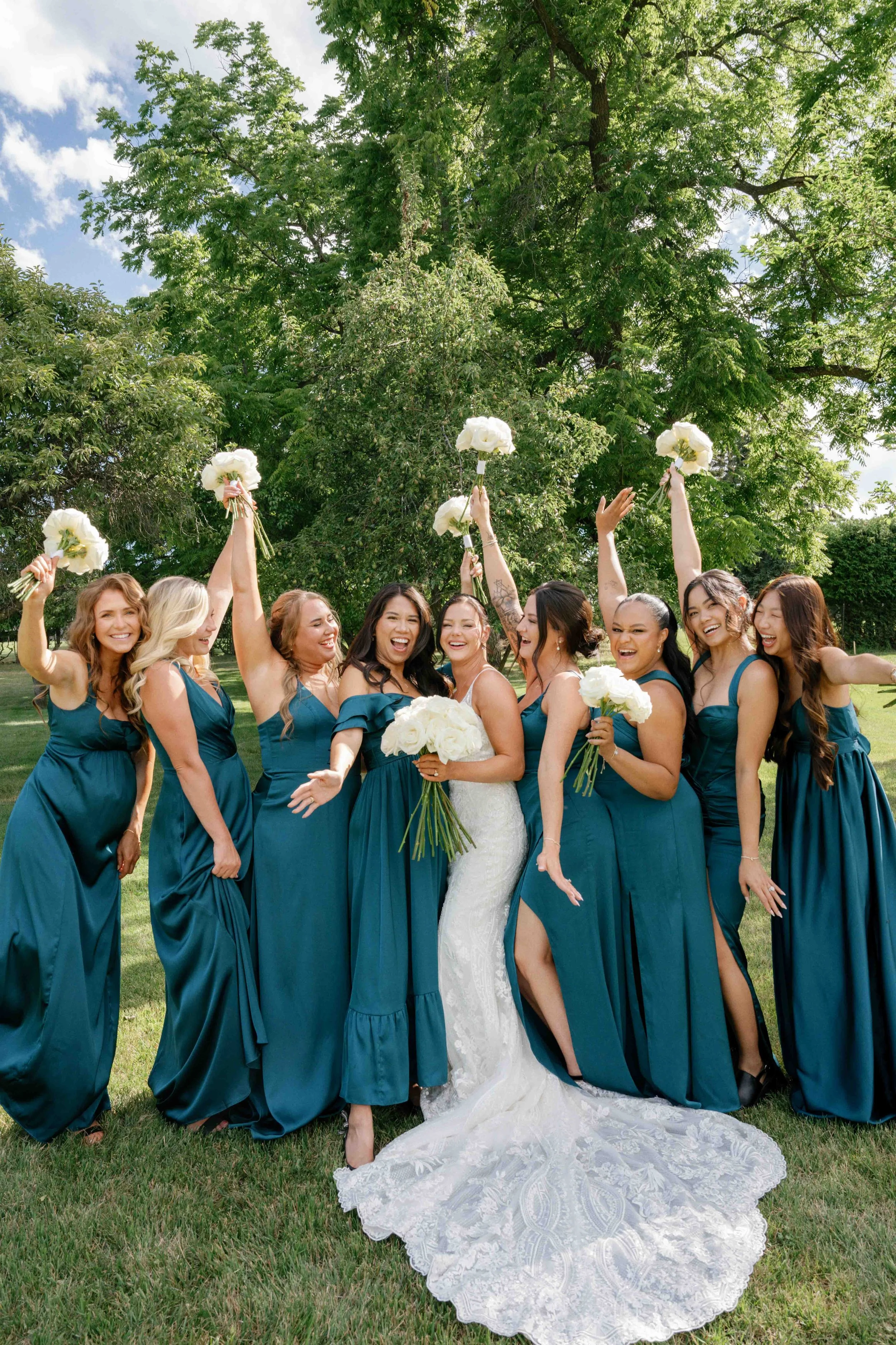 A group of women in teal dresses and one woman in a white wedding gown, holding white bouquets, standing on grass outdoors, celebrating a wedding.