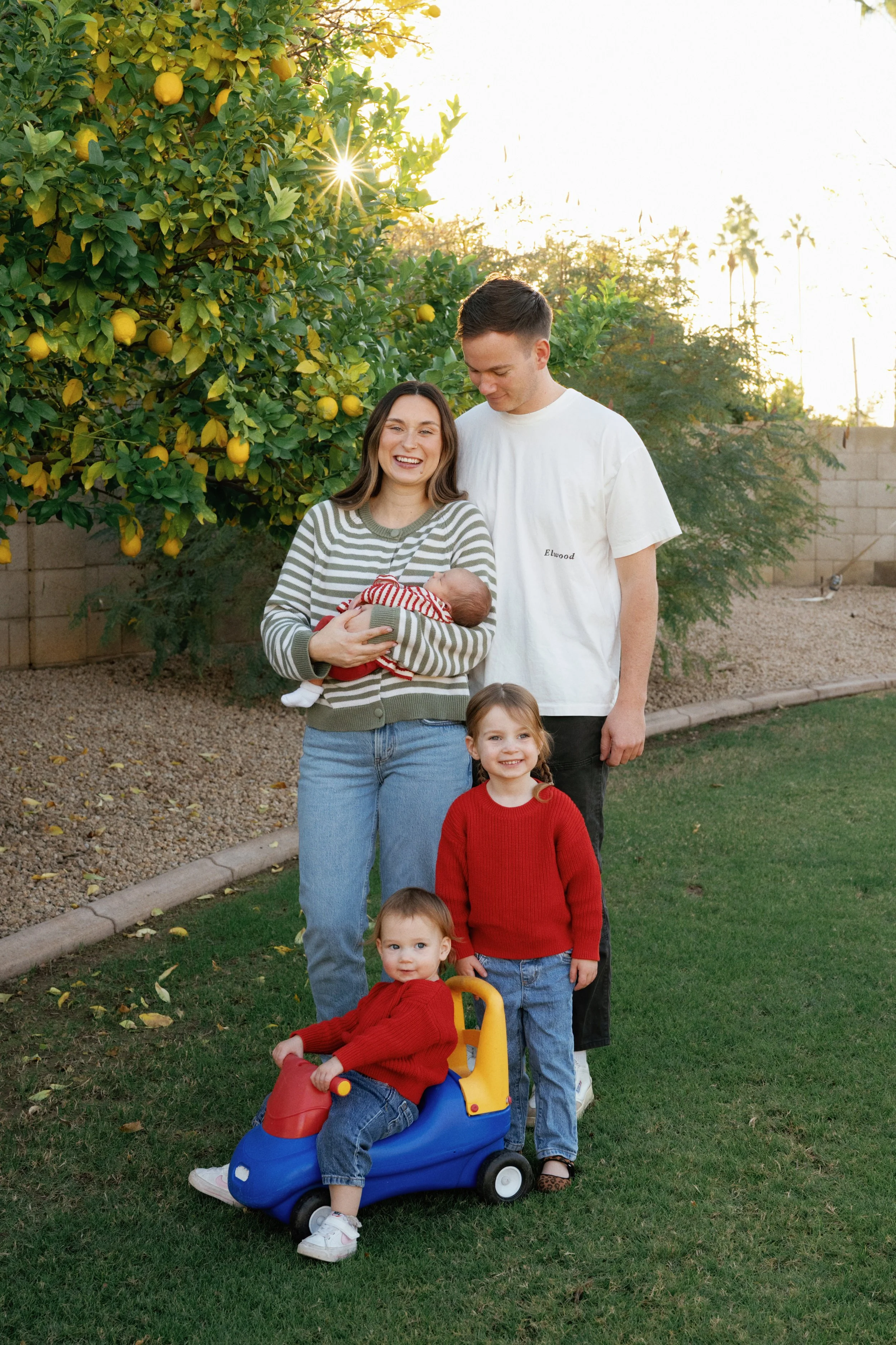 A family of five, including two children, a woman holding a baby, and a man, standing outdoors in a yard with lemon trees and a brick wall, during late afternoon sunlight. The children are smiling, and one boy is sitting on a blue and red ride-on toy