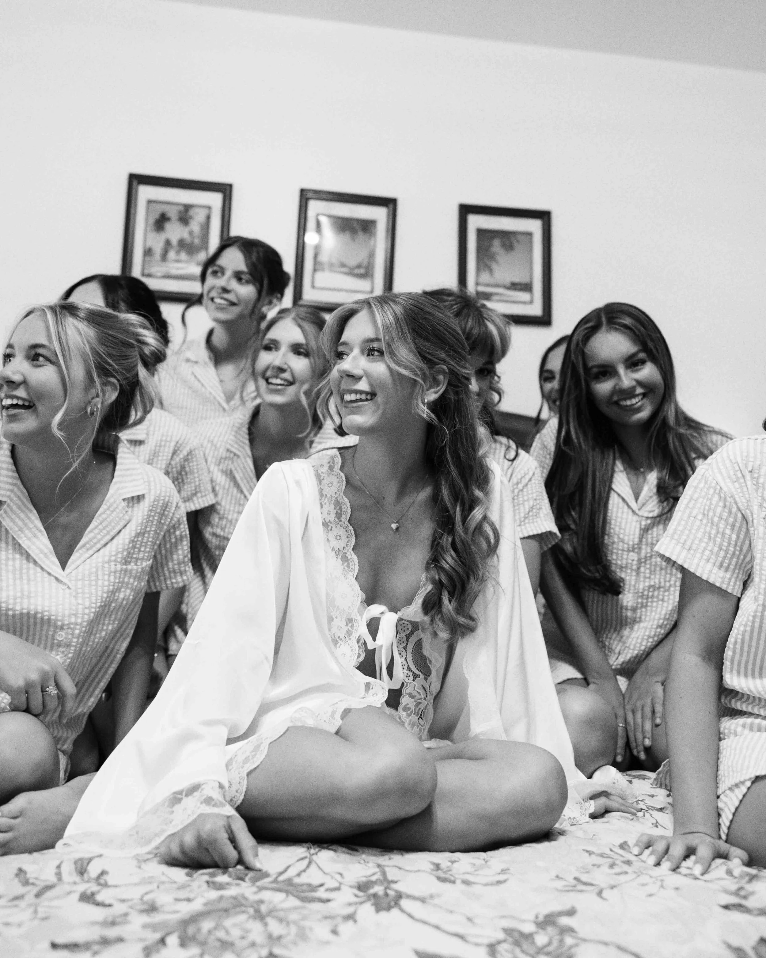 A group of young women sitting closely together indoors, smiling and looking in the same direction, with three framed pictures hanging on the wall behind them.