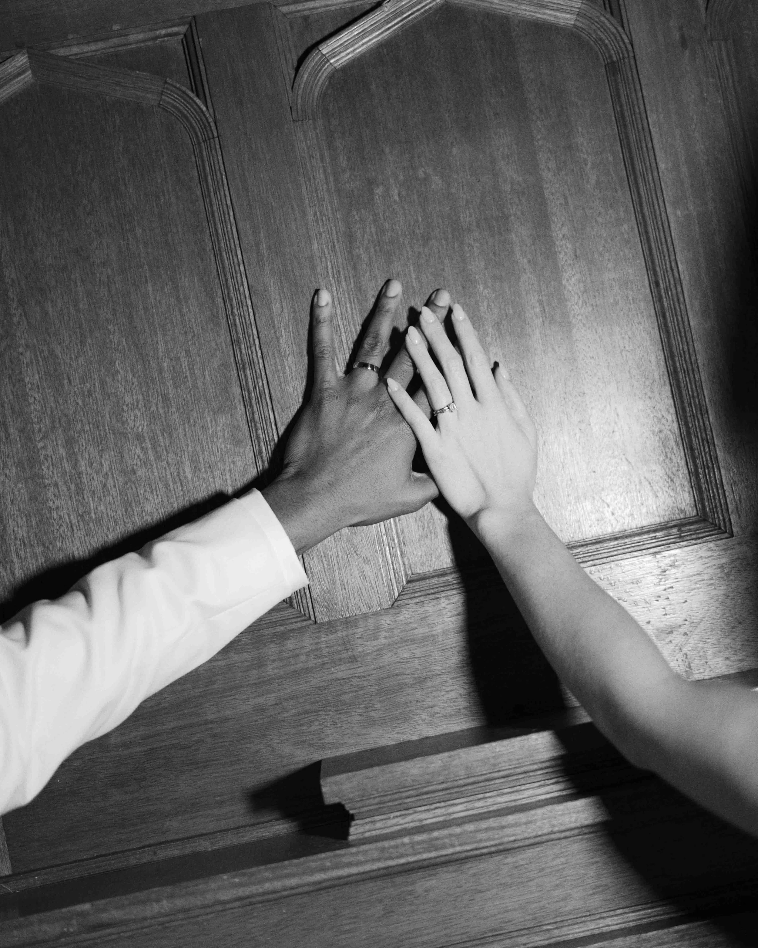 Two hands, one with a wedding ring, touching on a wooden table surface, black and white
