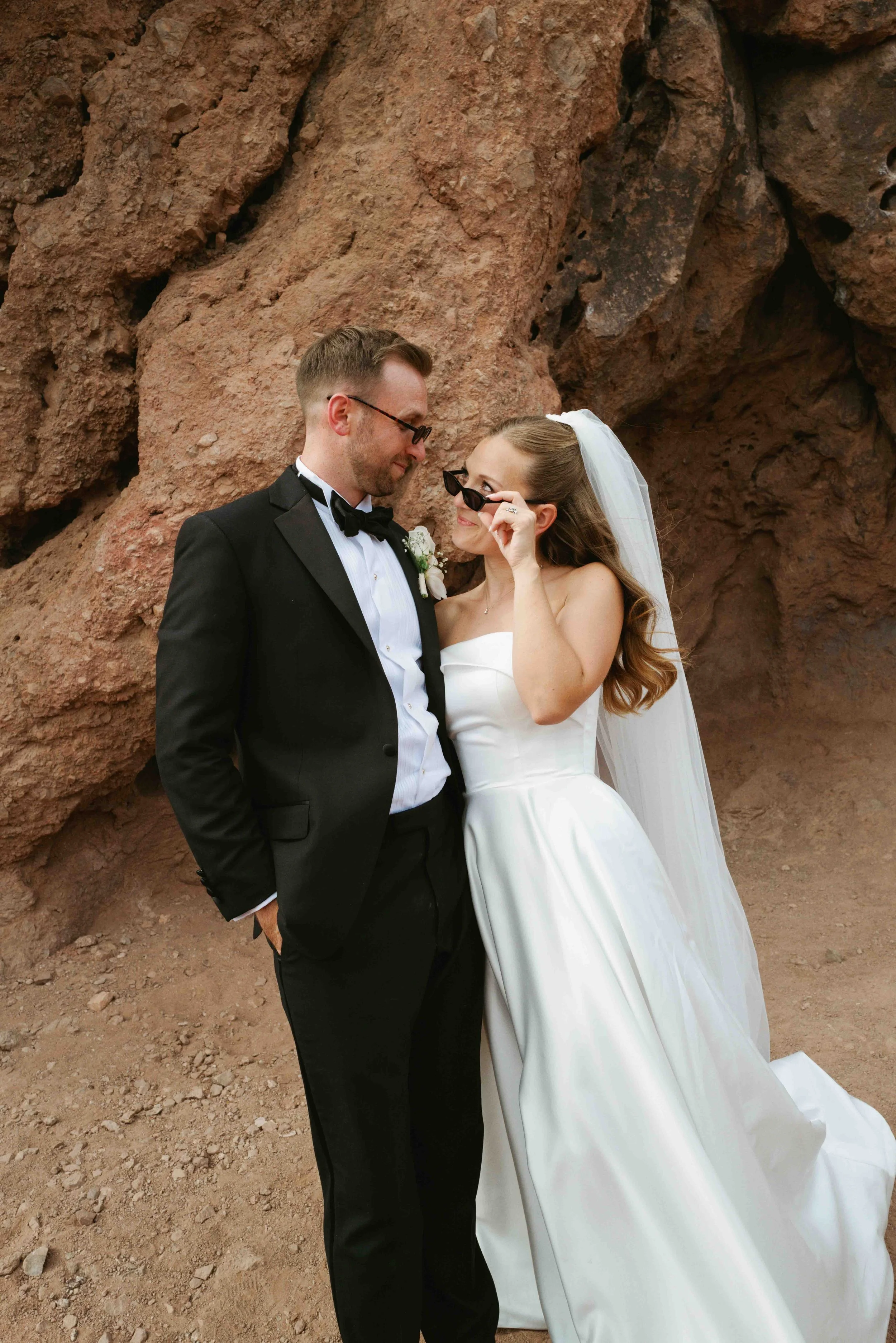 A bride and groom in wedding attire standing close together outdoors against a rocky background. The groom is dressed in a black tuxedo with a bow tie, and the bride is wearing a strapless white wedding gown with a veil. The bride is adjusting her su