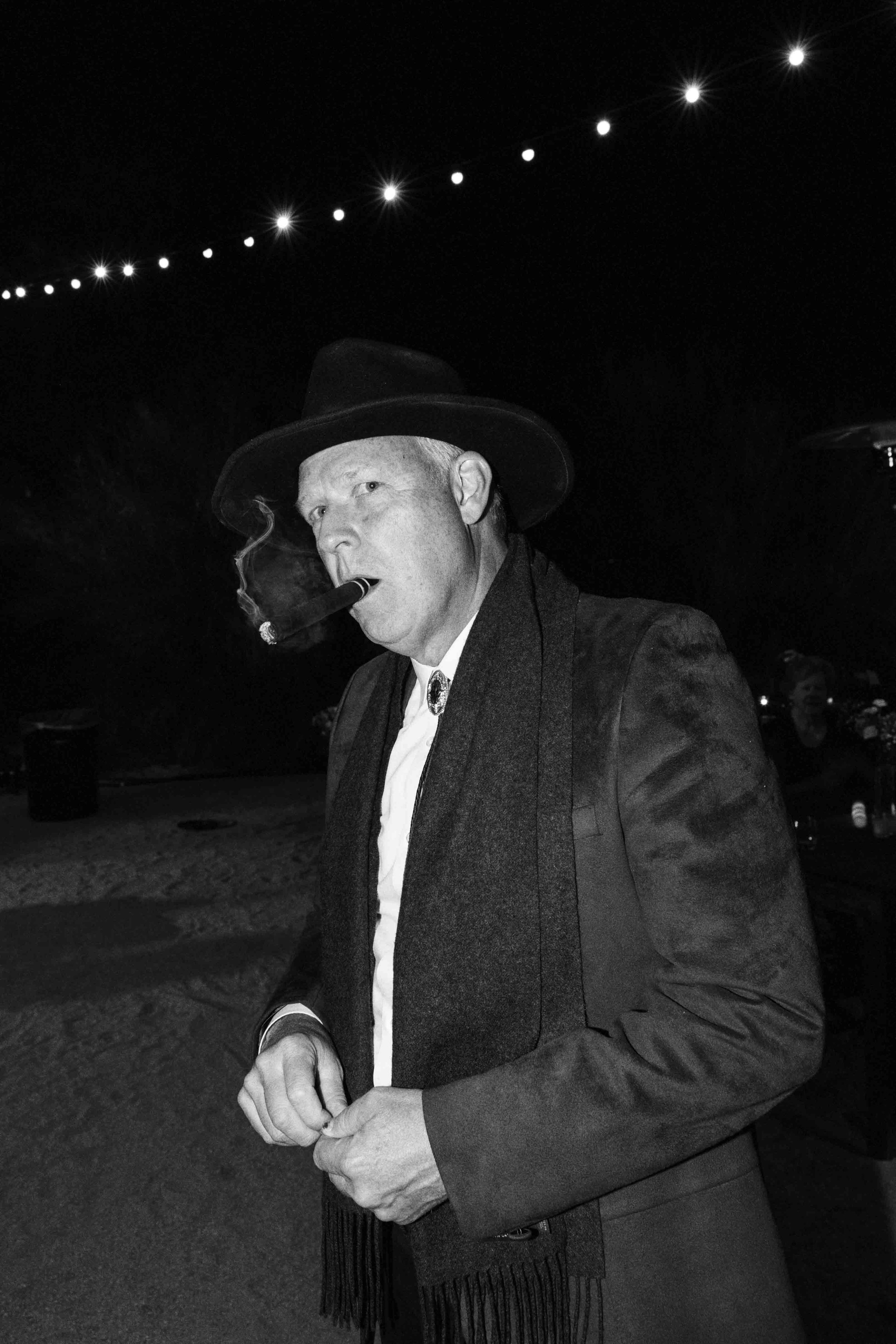 A man wearing a hat and dark coat smokes a cigar in a dark outdoor setting, with string lights visible in the background.