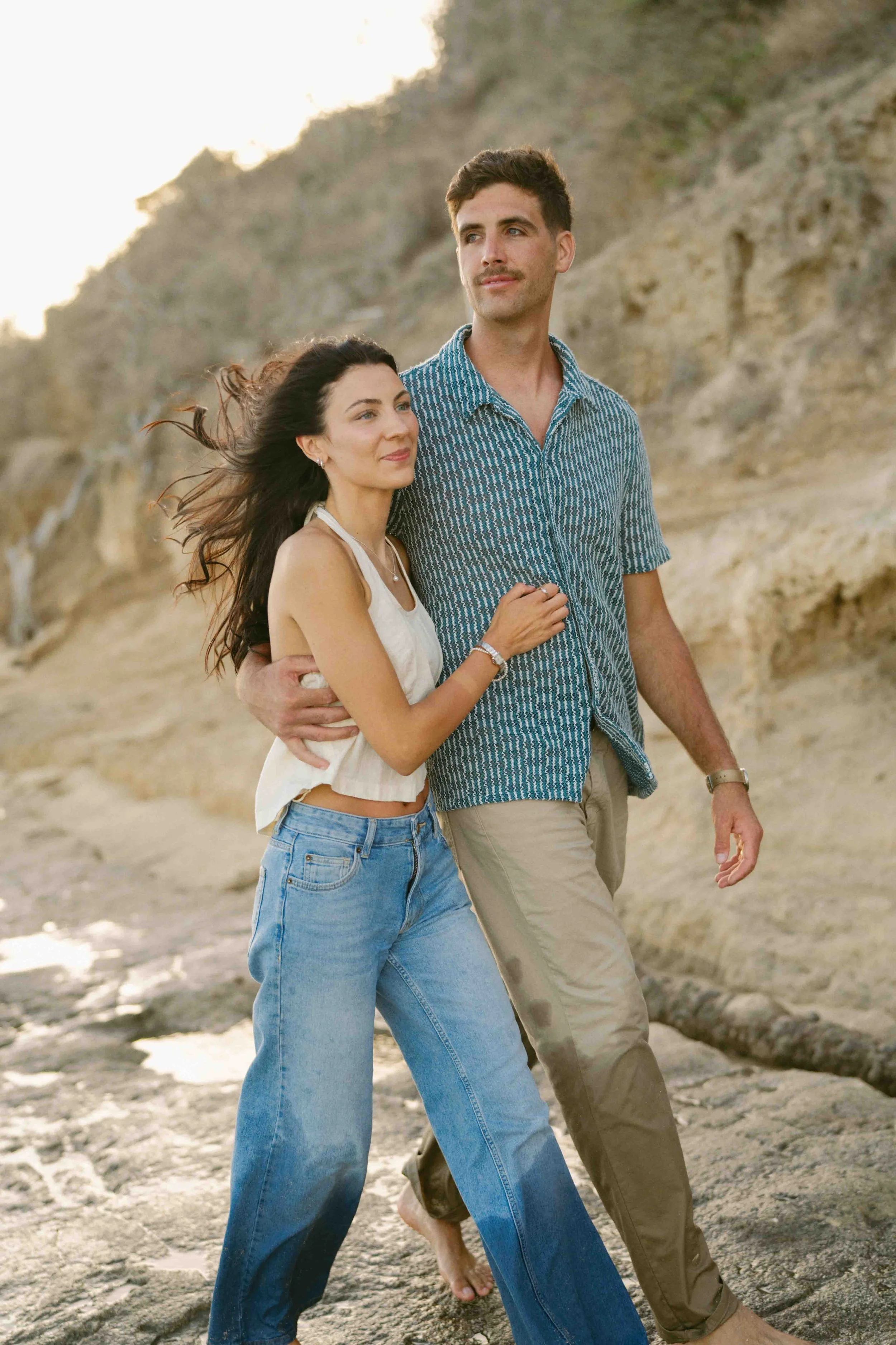 A smiling woman with dark hair and a man with short hair and a mustache walking on the beach during sunset, with rocky cliffs in the background.
