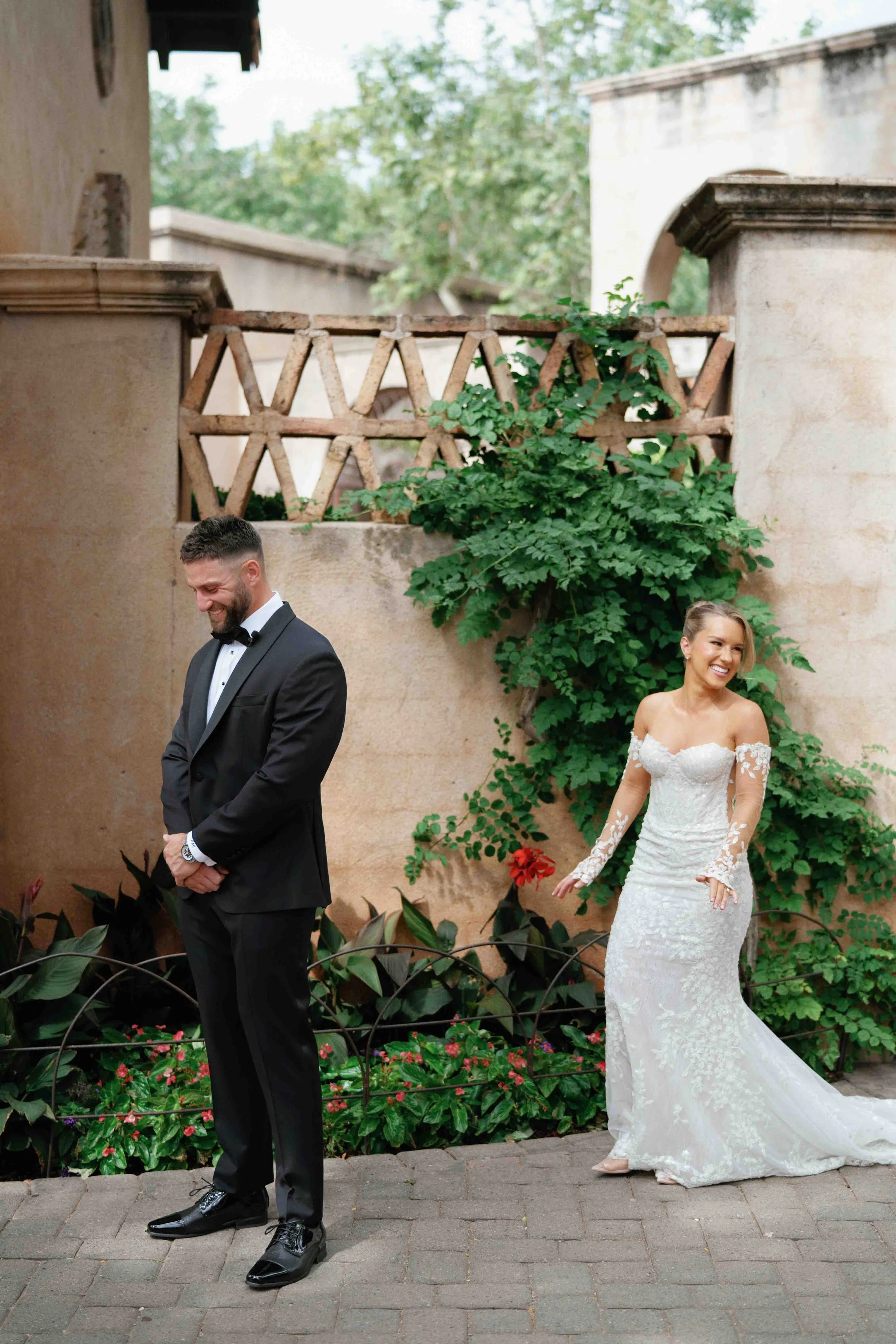 A man in a black tuxedo with a bow tie is standing with his head bowed and eyes closed. A woman in a white wedding dress with long lace sleeves and floral lace detail is smiling and standing behind him. They are outdoors near a beige stone wall with 